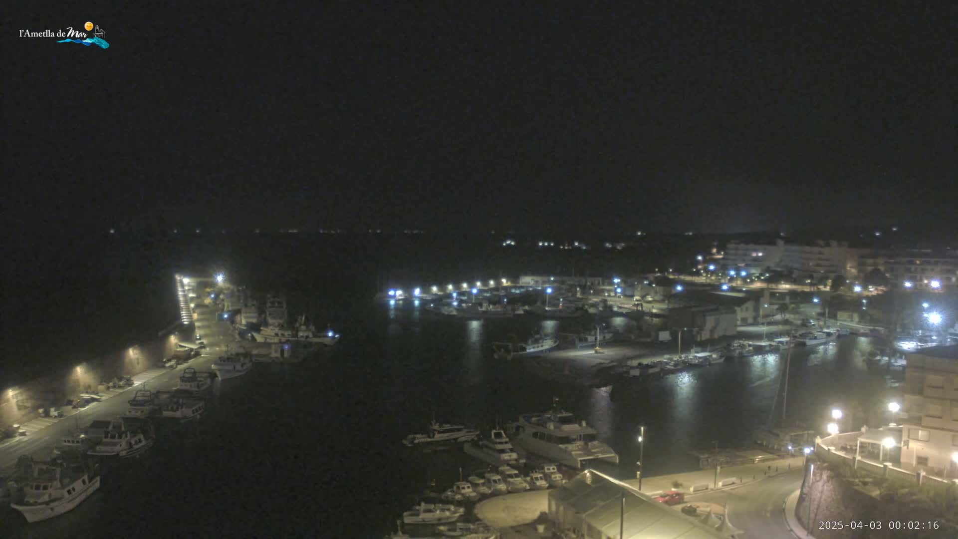 A nighttime aerial view of a harbor filled with boats, illuminated by streetlights and the lights of buildings on the shoreline.
