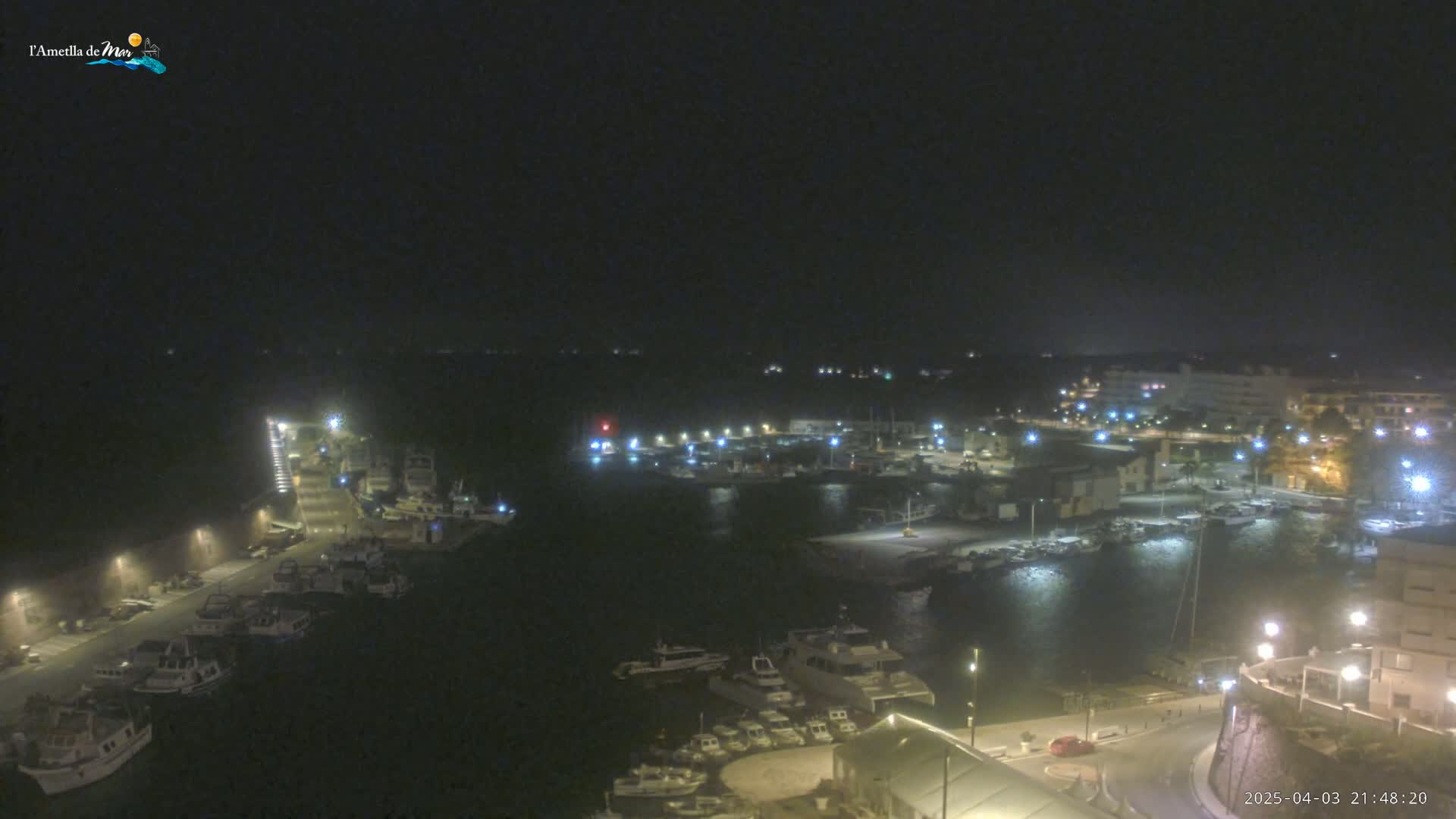 A nighttime, aerial view shows a harbor filled with boats and surrounded by buildings with illuminated streets.