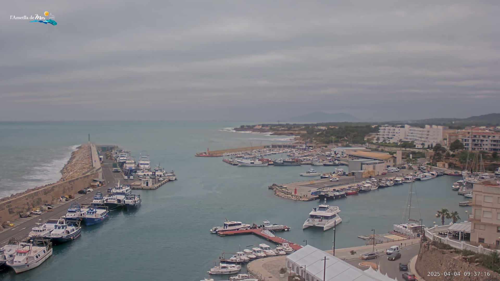 A harbor filled with numerous boats is shown under an overcast sky, with waves crashing against a breakwater.