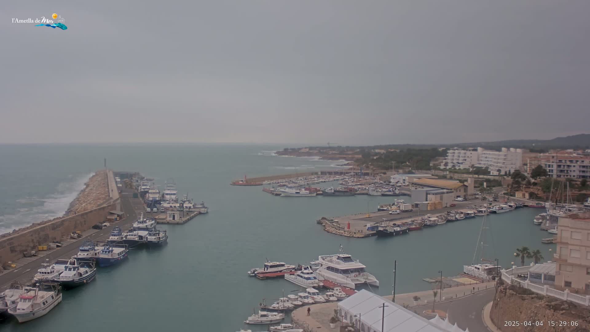 A harbor filled with numerous boats is shown on an overcast day, with waves crashing against a breakwater to the left.
