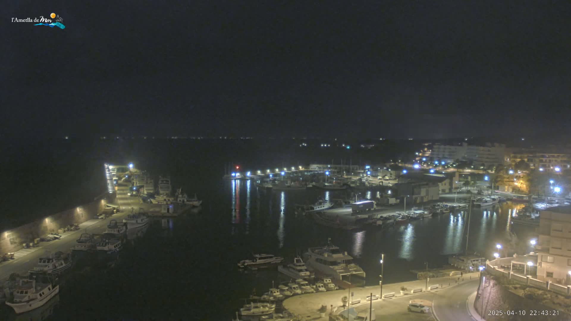 A nighttime view of a harbor filled with boats, illuminated by lights from the surrounding buildings and docks under a dark sky.