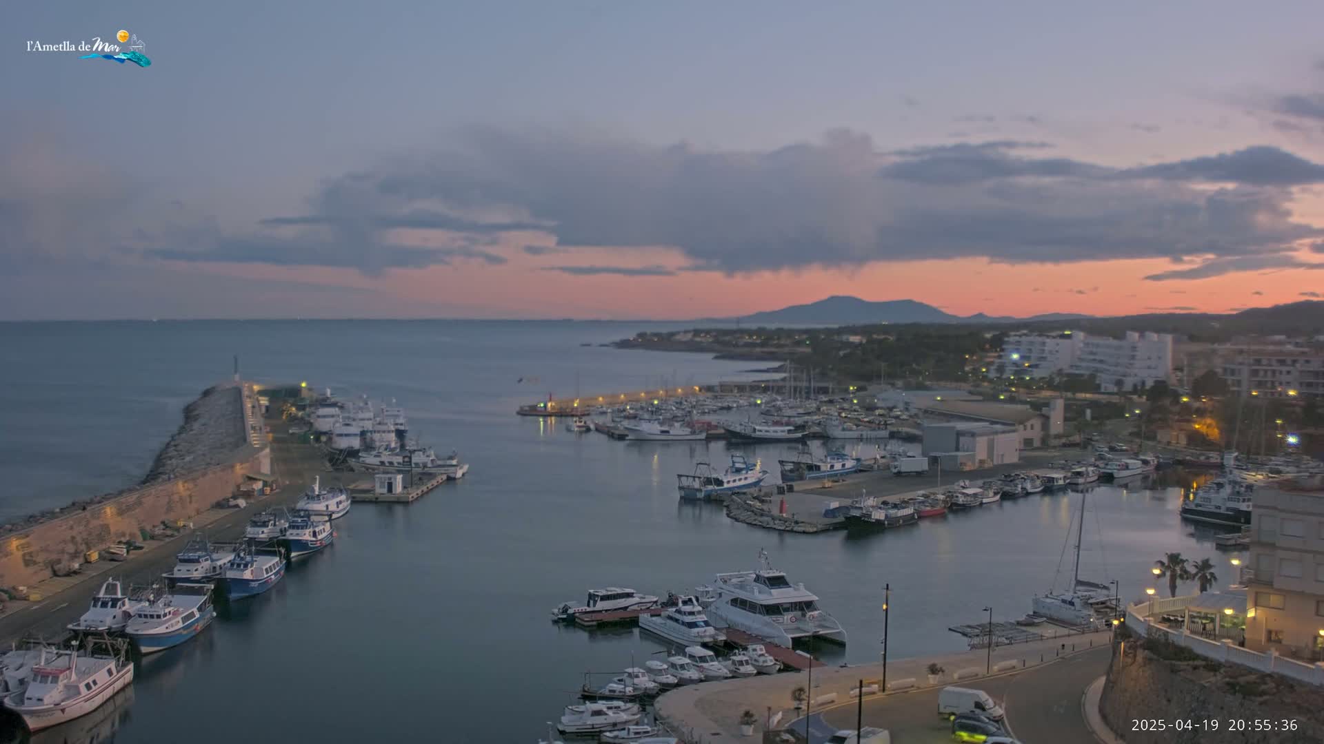 A harbor filled with numerous boats is shown at dusk under a partly cloudy sky with a pinkish hue on the horizon.