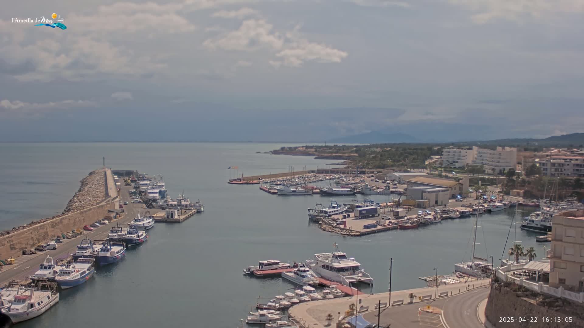 A harbor filled with numerous boats and ships under a partly cloudy sky, is situated next to a town with buildings and roads.