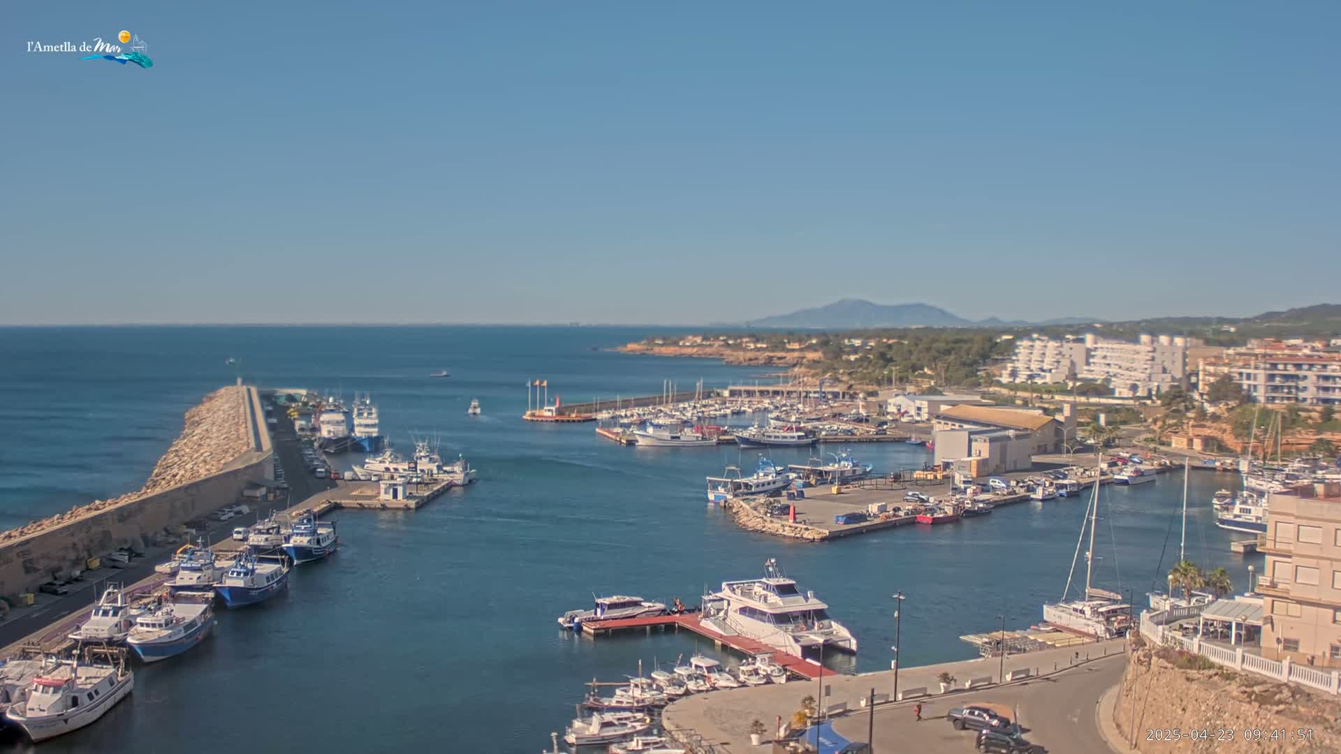 A harbor filled with numerous boats and yachts is situated along a calm, clear ocean under a bright, sunny sky.