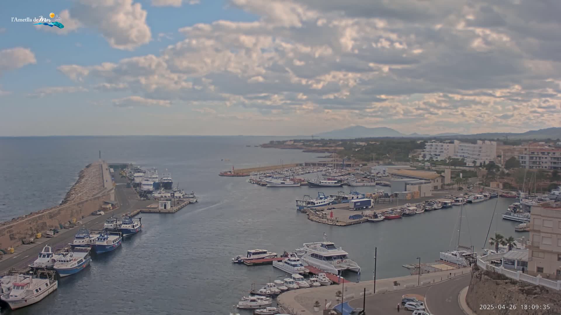 A partly cloudy day overlooks a harbor filled with many boats of various sizes, some docked and others in the water, with a stone breakwater extending into the sea.