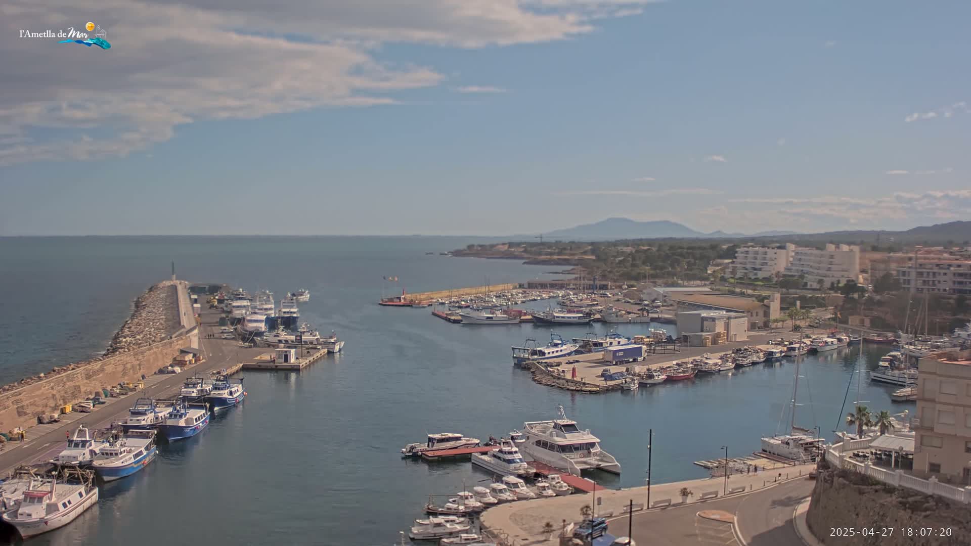 A harbor filled with numerous boats and yachts is shown under partly cloudy skies, with a stone breakwater extending into the calm sea.