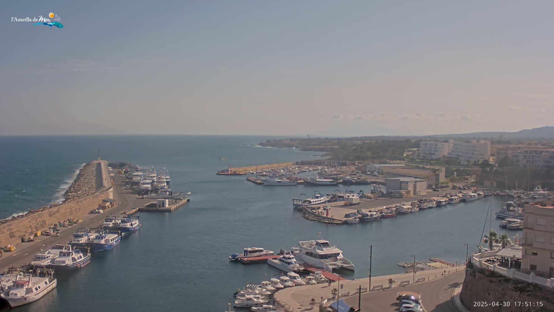 A harbor filled with numerous boats and ships is seen on a sunny day, with calm water and a rocky breakwater extending into the sea.