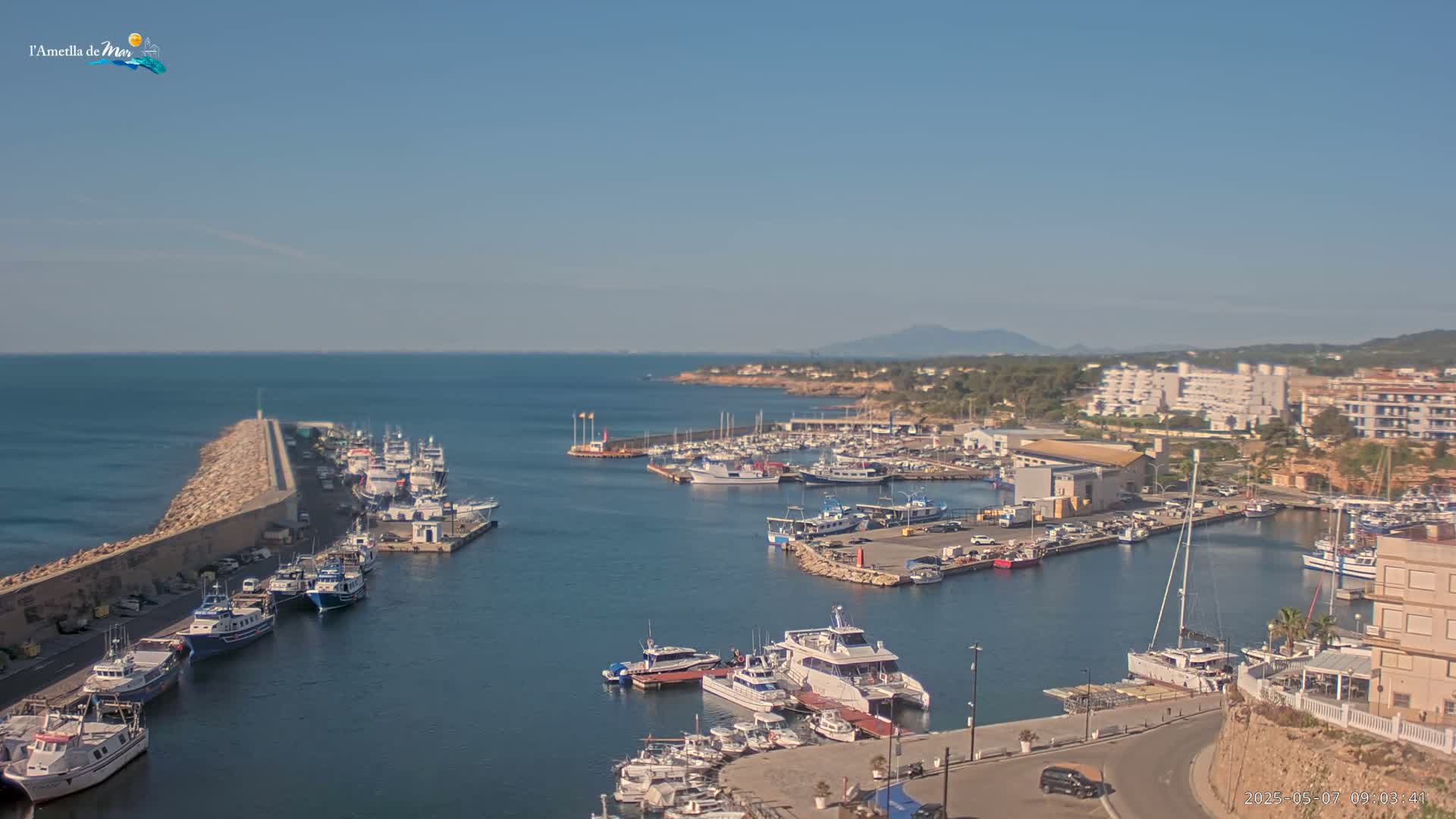A harbor filled with numerous boats and yachts is situated next to a stone wall that extends into a calm, clear blue sea under a sunny sky.