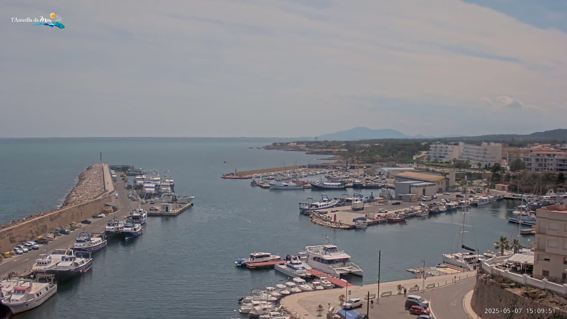 A harbor filled with numerous boats and yachts is seen on a partly cloudy day, with mountains visible in the distance.