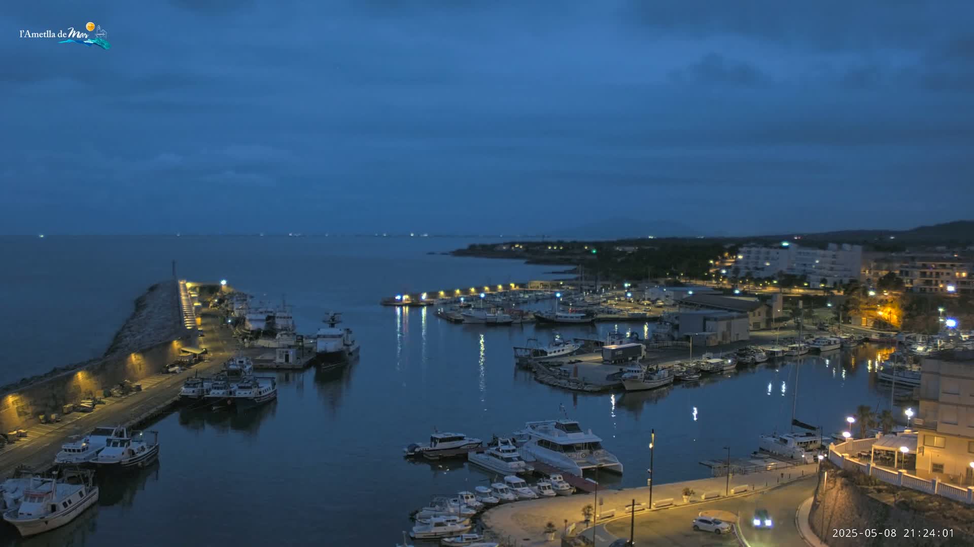 A nighttime view of a harbor filled with boats, illuminated by lights, under a dark blue, cloudy sky.