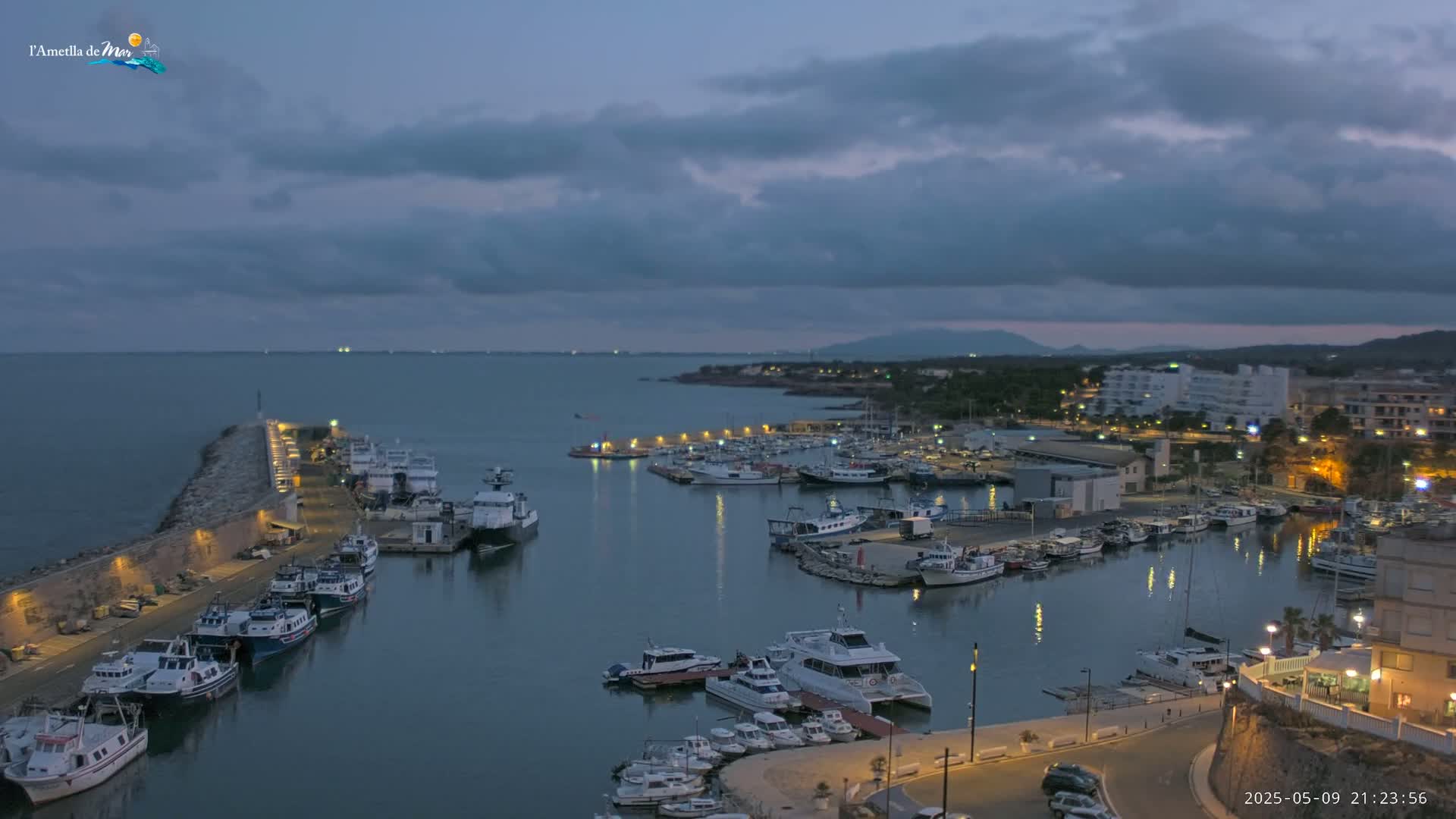 A harbor filled with numerous boats is nestled along a coastline under a cloudy twilight sky.