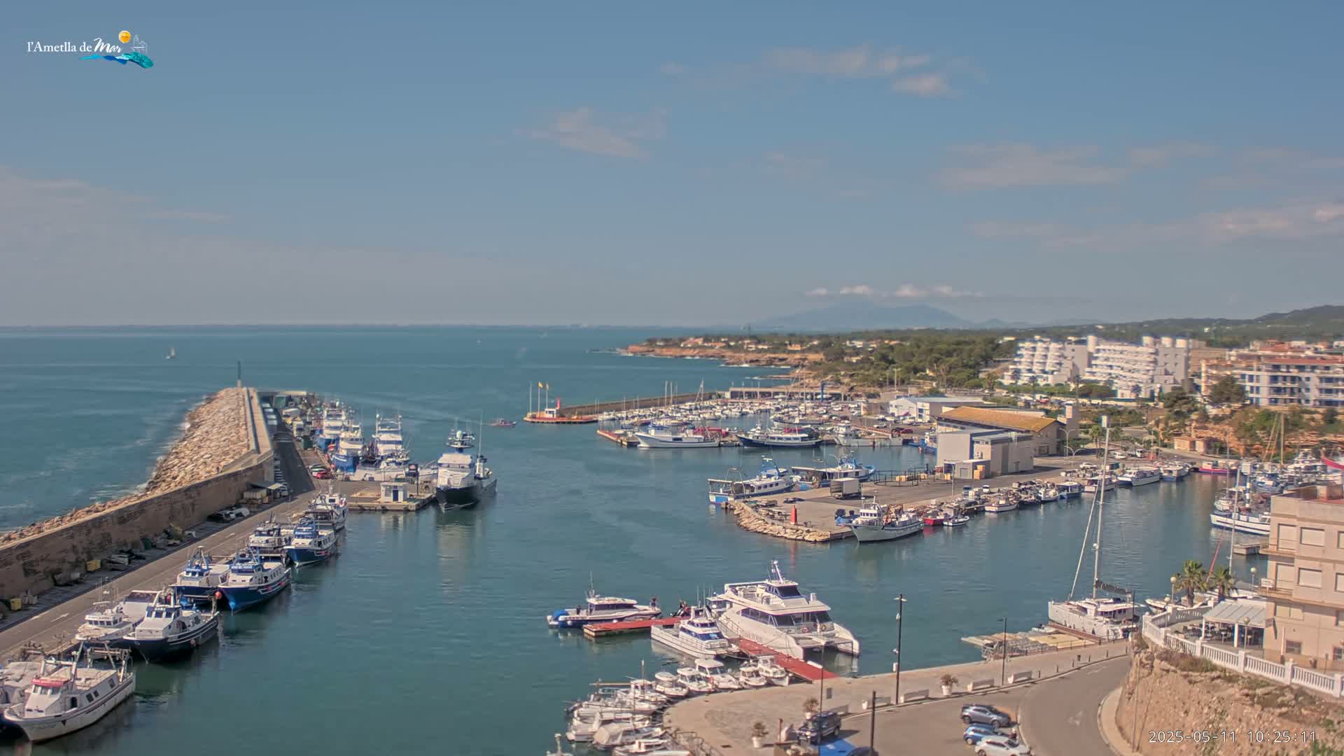 A harbor filled with numerous boats and yachts is situated along a coastline under a mostly sunny sky.