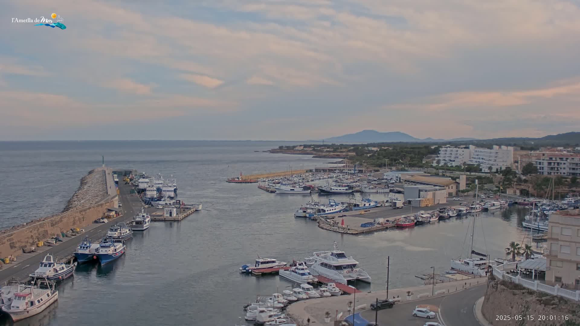 A harbor filled with numerous boats and yachts is shown under a partly cloudy sky, with buildings and a coastline visible in the background.