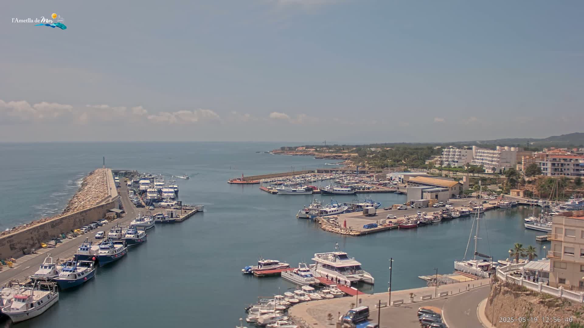 A mostly sunny day reveals a harbor filled with numerous boats, some docked and others in the water, with buildings and a stone breakwater visible in the background.