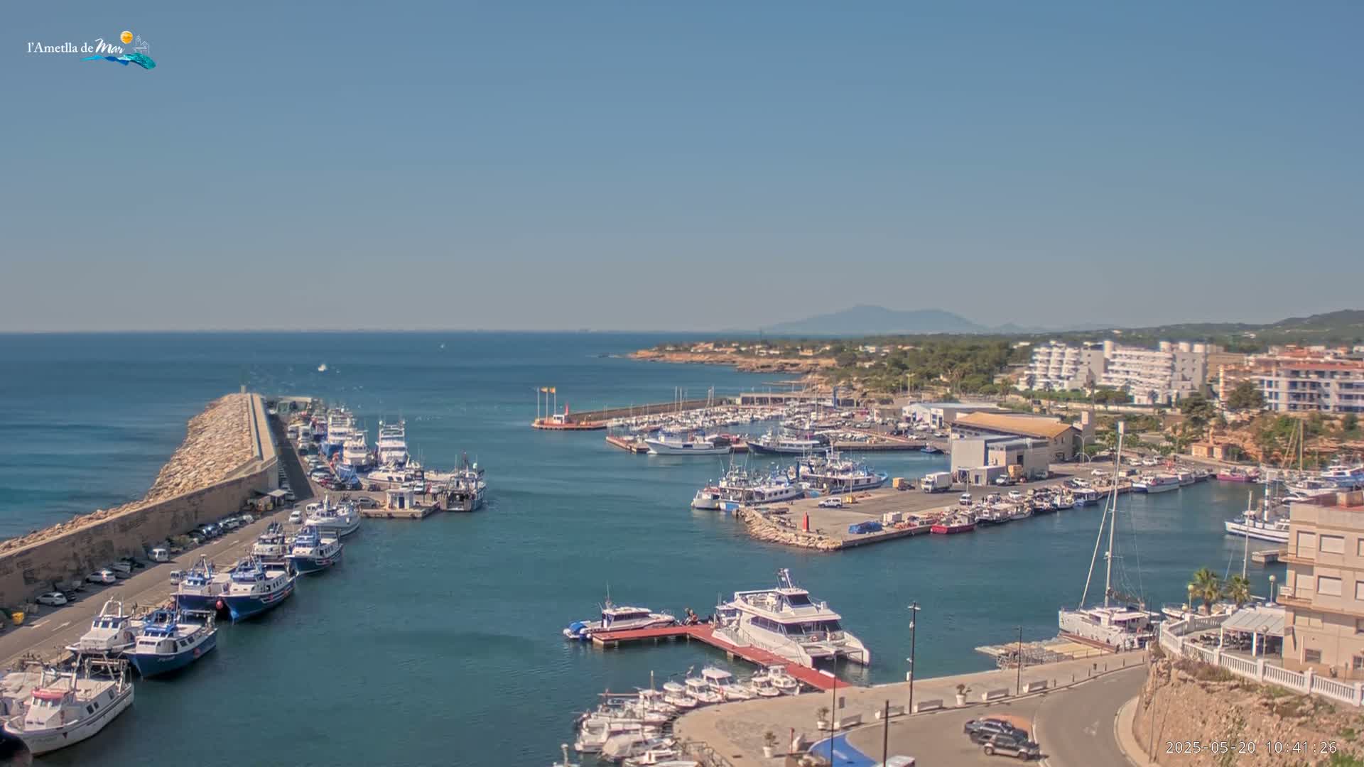 A harbor filled with numerous boats and yachts is seen on a sunny day, with calm blue water and a distant coastline under clear skies.