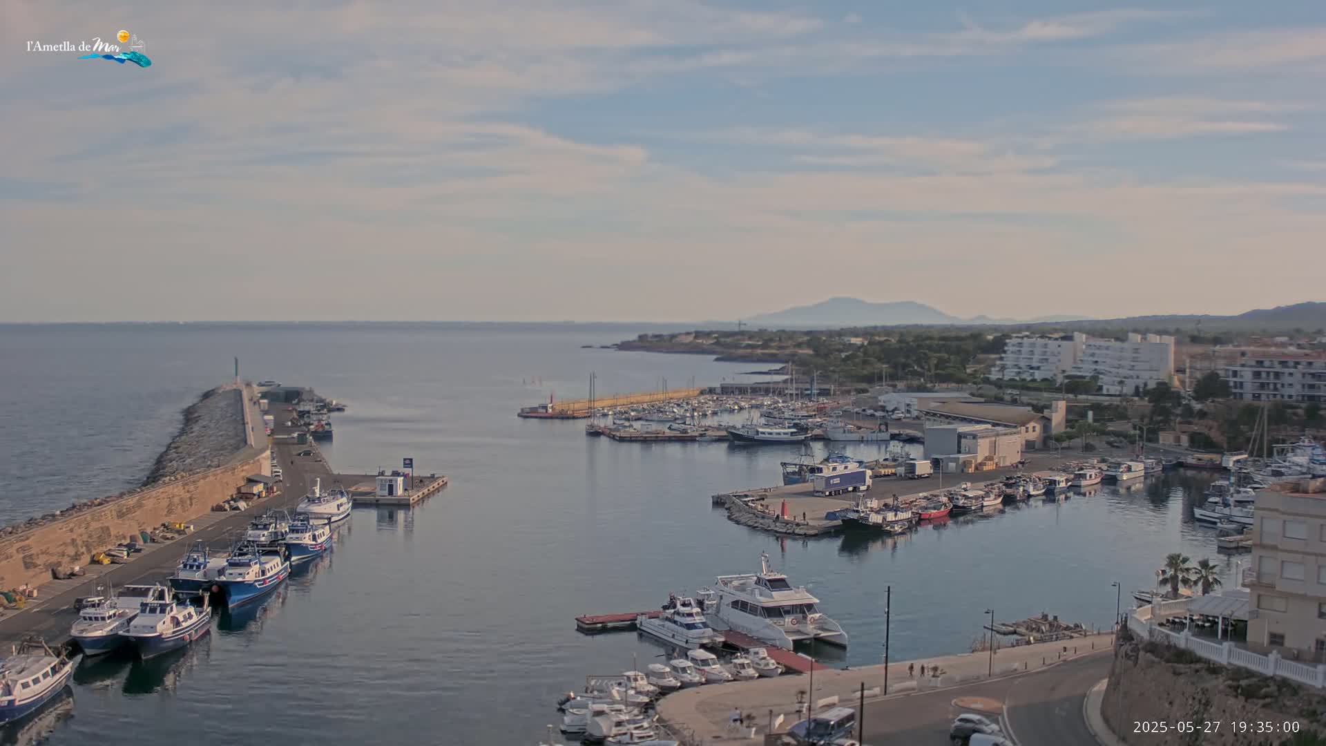 A harbor filled with numerous boats and yachts is situated beside a stone breakwater, under a partly cloudy sky.