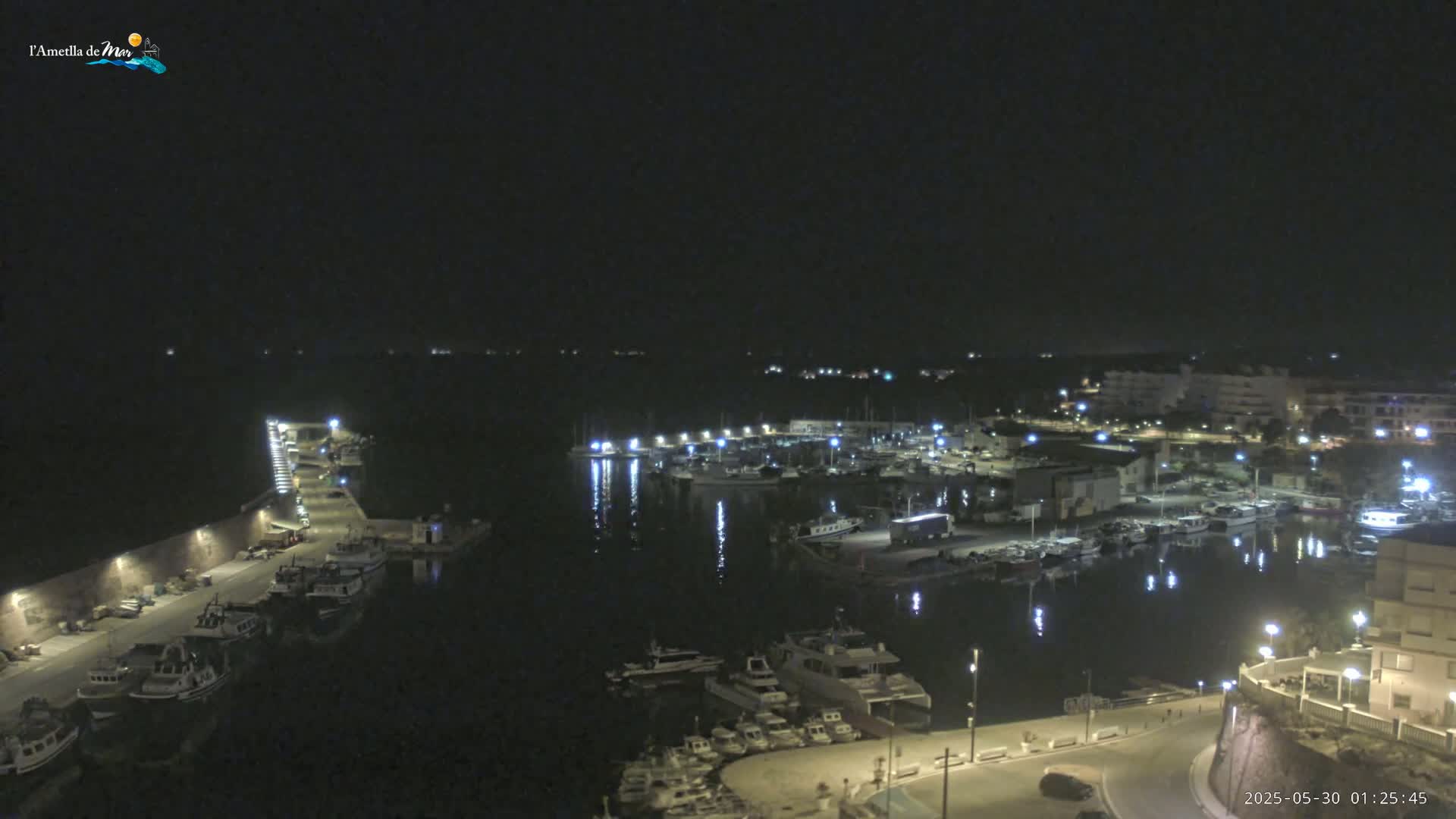 A nighttime aerial view of a harbor filled with boats, illuminated by lights from buildings and street lamps along a waterfront road.