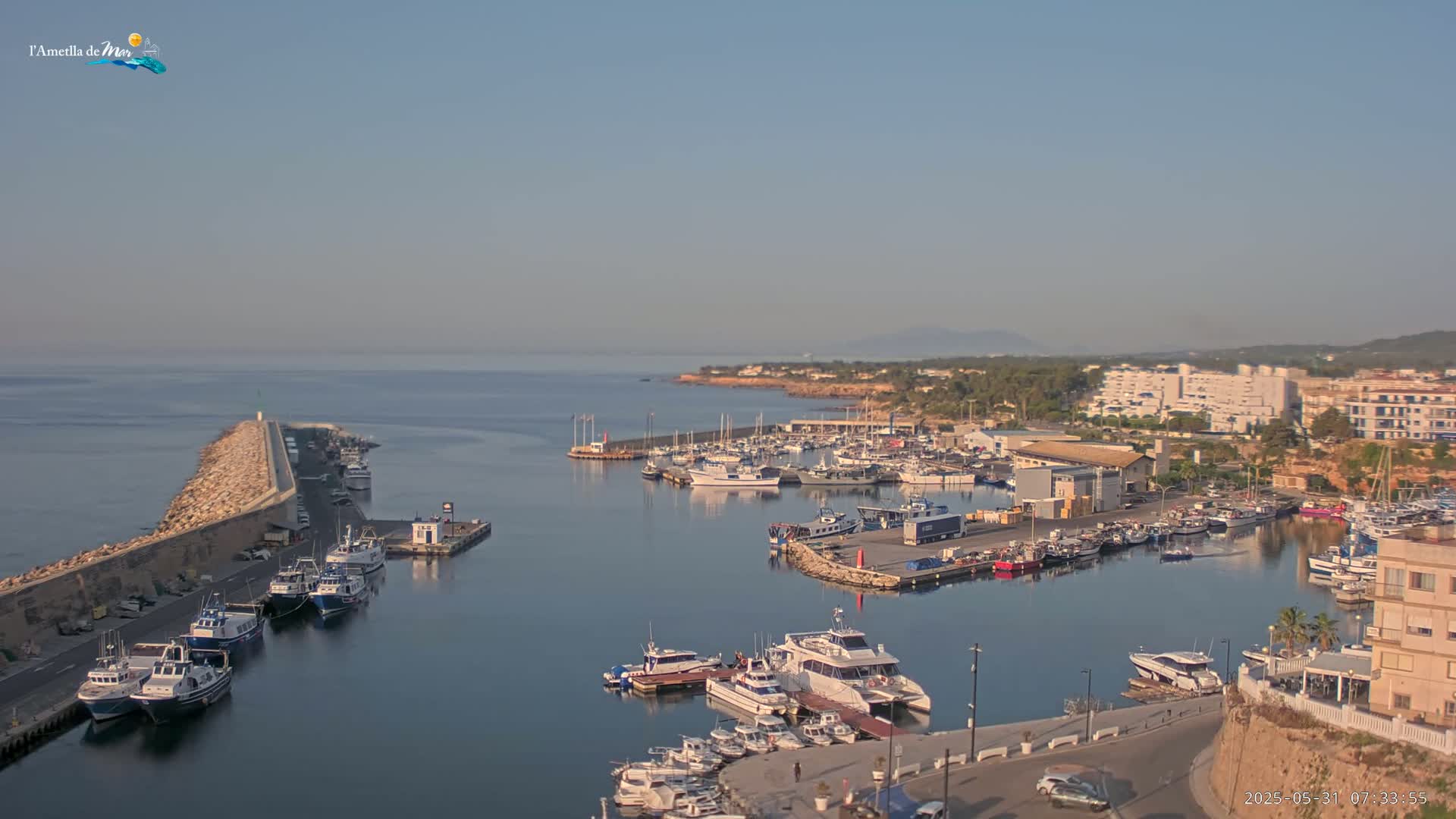 A harbor filled with numerous boats and yachts is seen on a clear, sunny day, with a stone breakwater extending into the calm sea.
