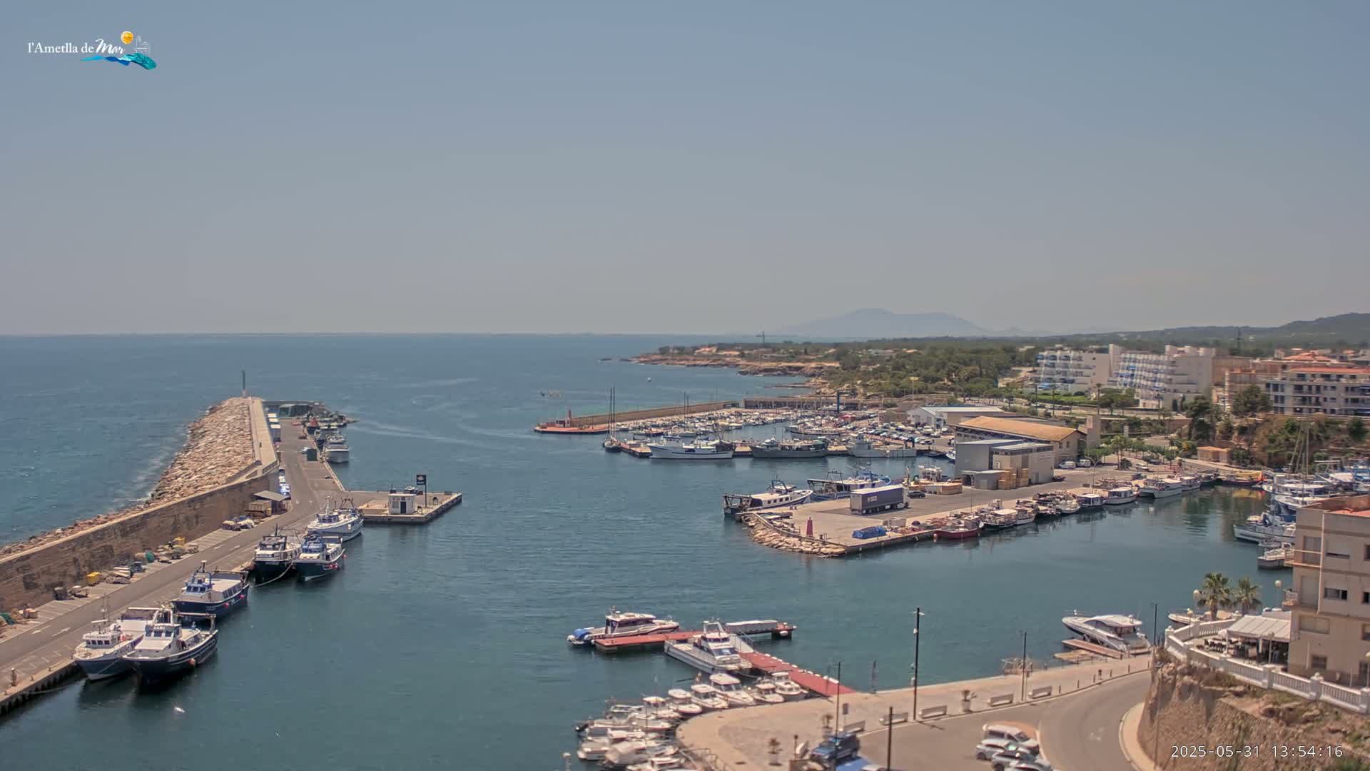 A harbor filled with numerous boats is shown on a sunny day, with a stone breakwater extending into a calm sea.