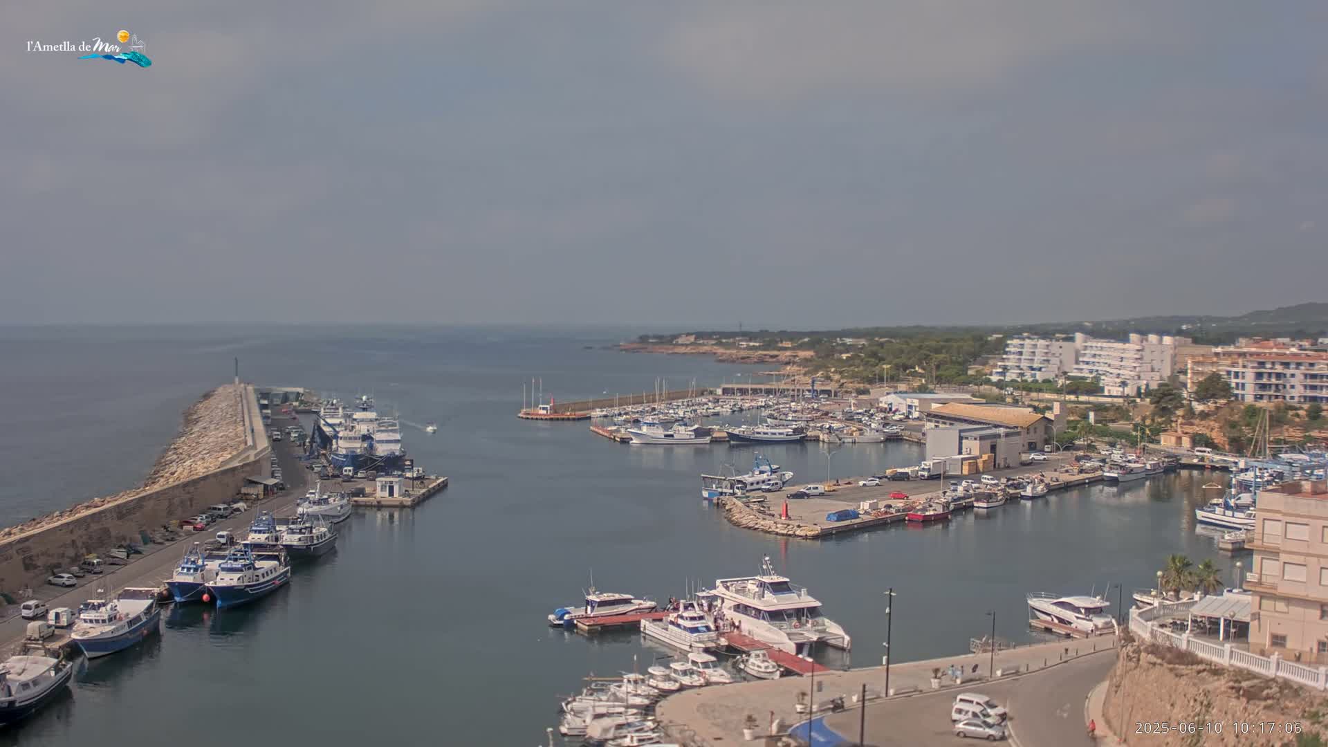 A harbor filled with numerous boats and yachts is seen on a partly cloudy day, with a stone breakwater extending into the calm sea.