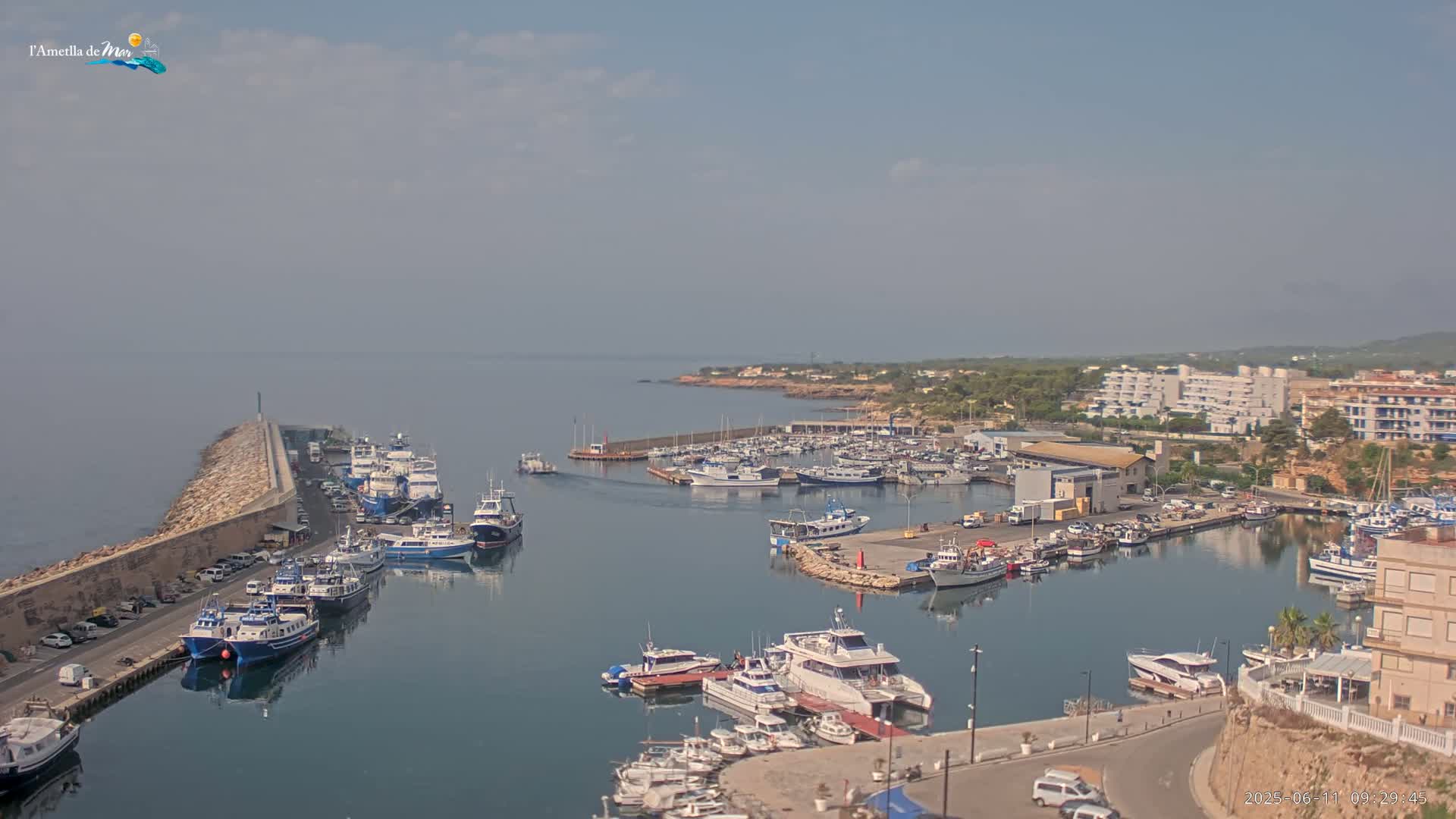 A hazy day reveals a harbor filled with numerous boats and yachts, alongside a stone seawall and coastal buildings.