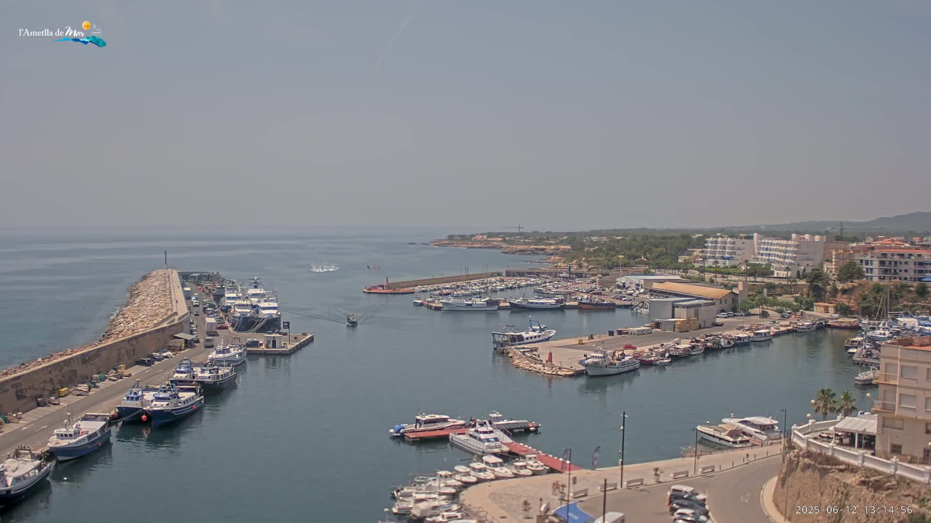 A harbor filled with numerous boats and yachts is seen on a hazy day, nestled against a coastline with buildings and vegetation.