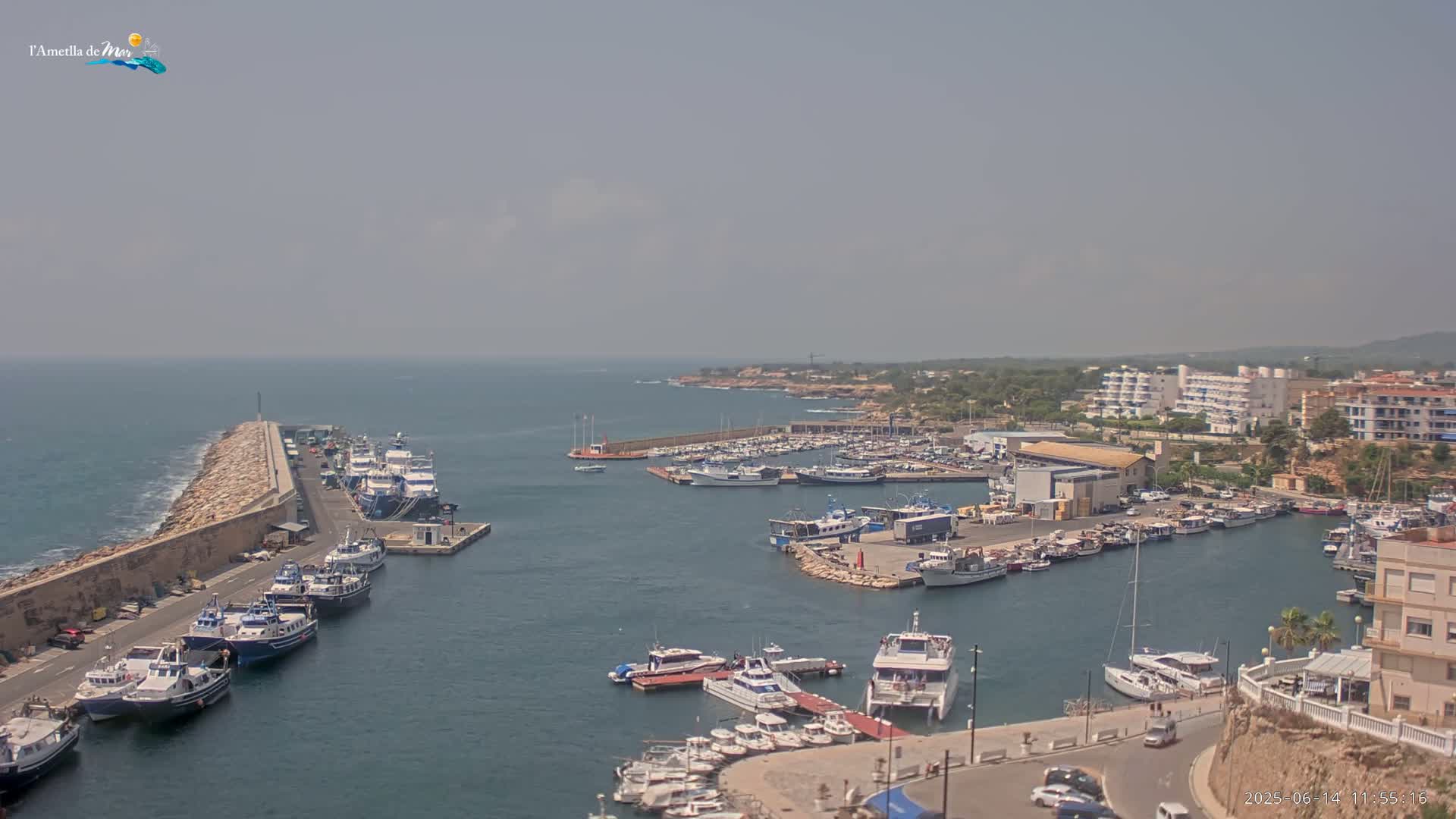 A hazy day reveals a harbor filled with numerous boats and yachts, nestled alongside a coastline featuring buildings and a stone breakwater.