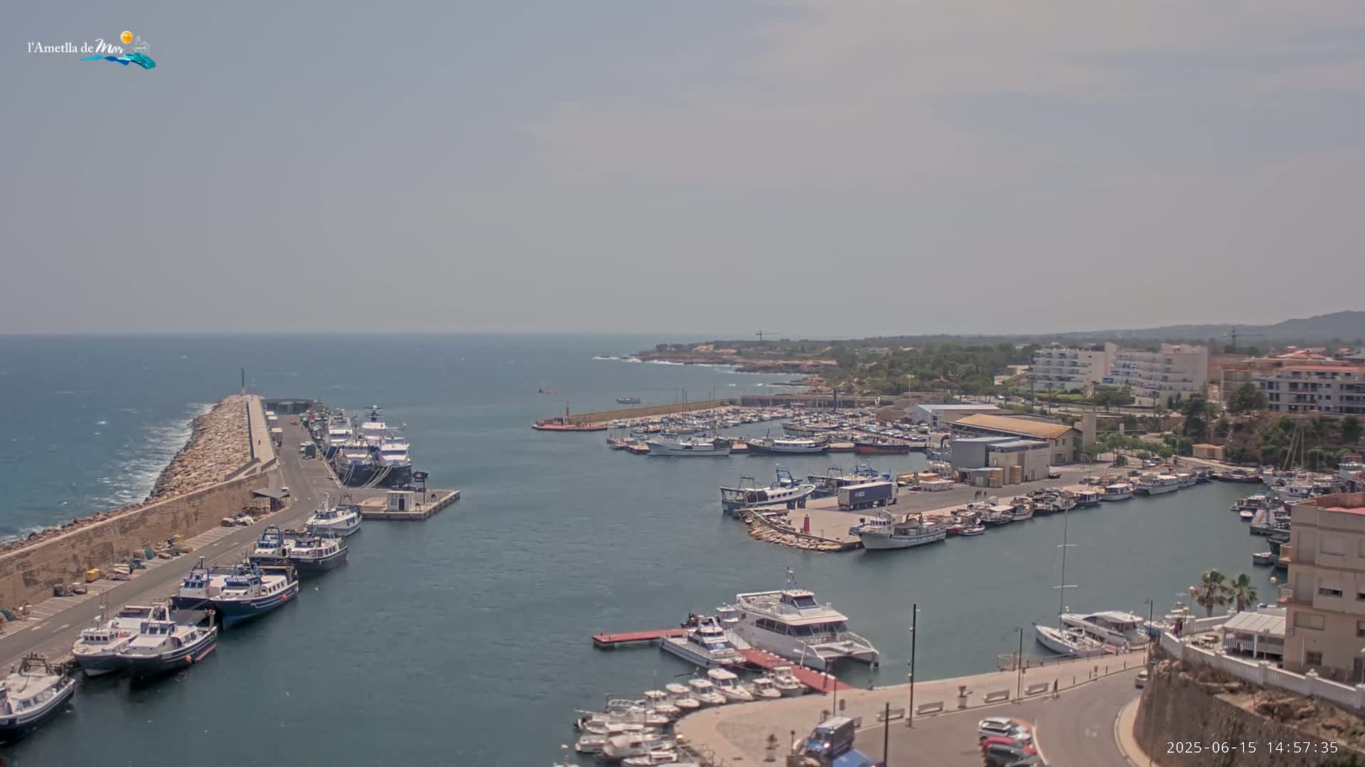 A harbor filled with numerous boats and yachts is shown on a hazy day, with a stone breakwater extending into a calm sea.