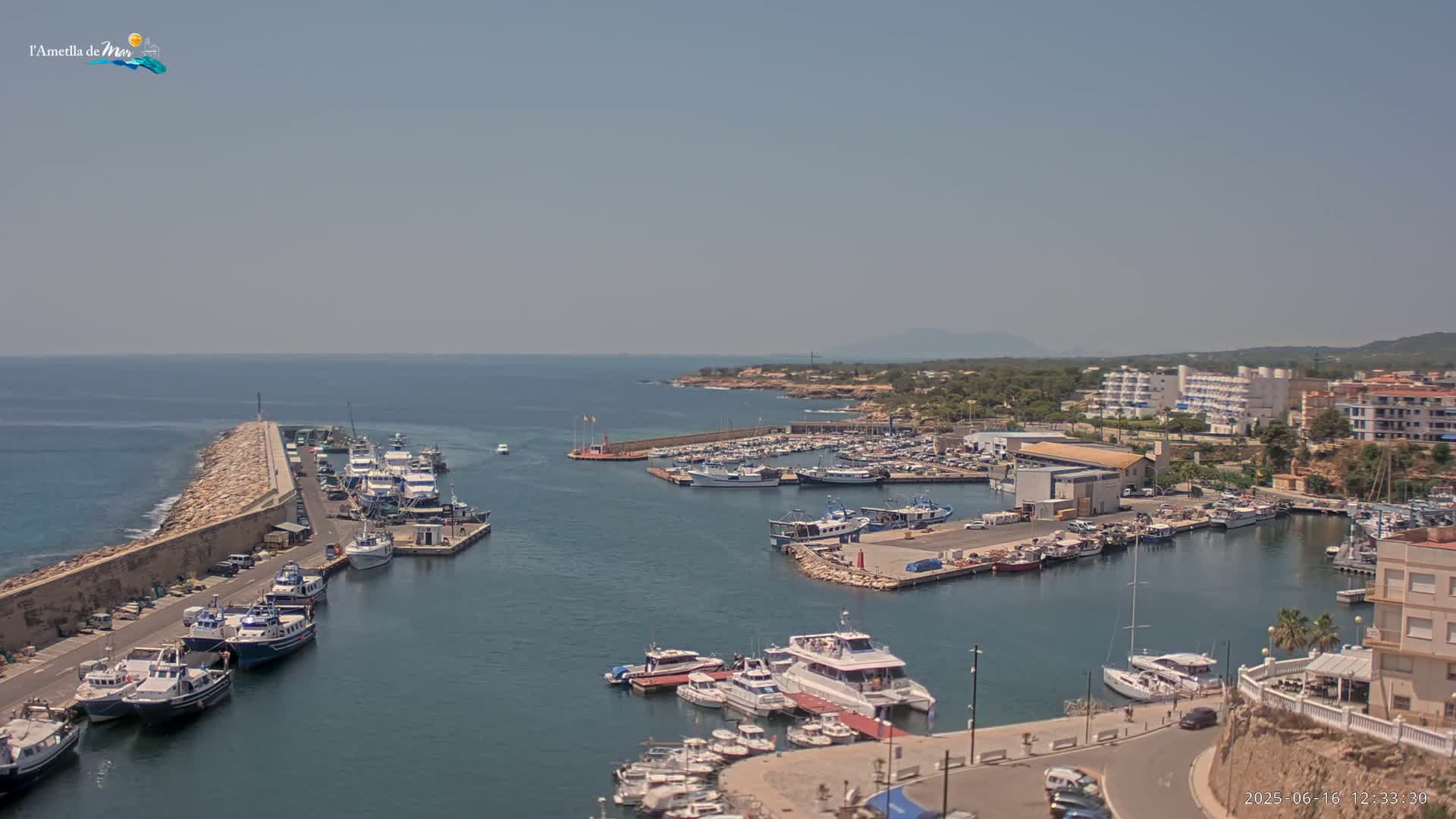 A sunny day shows a harbor filled with many boats, some docked and others in the water, situated next to a town with buildings along the coast.