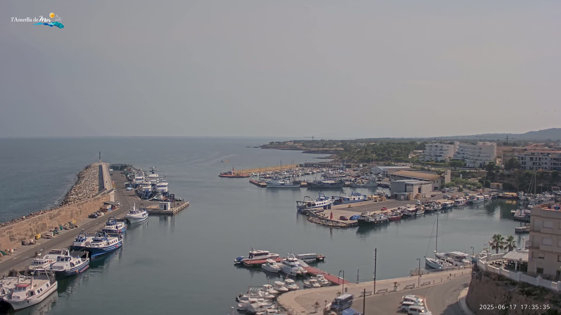 A harbor filled with numerous boats and yachts is seen on a hazy day, with a stone breakwater extending into the calm sea.
