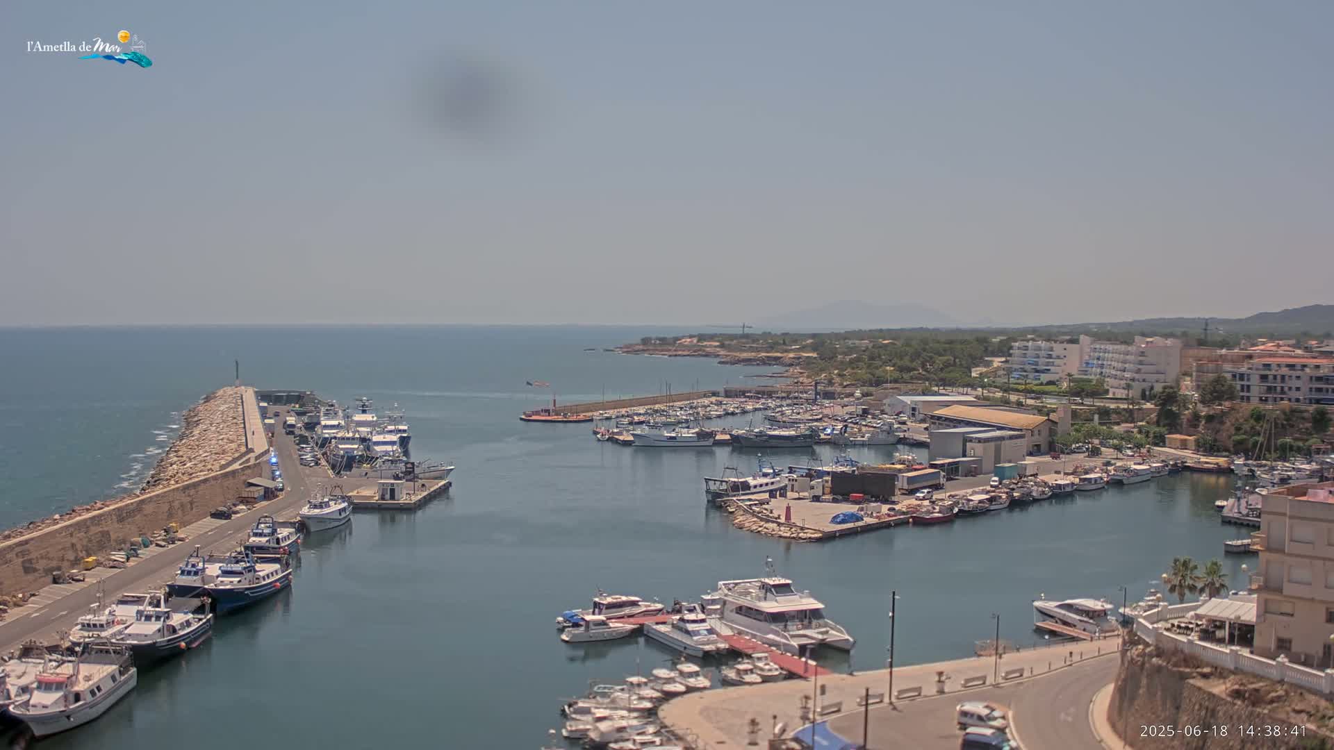 A harbor filled with numerous boats and yachts is situated beside a town on a sunny day, with calm waters extending to a distant coastline.