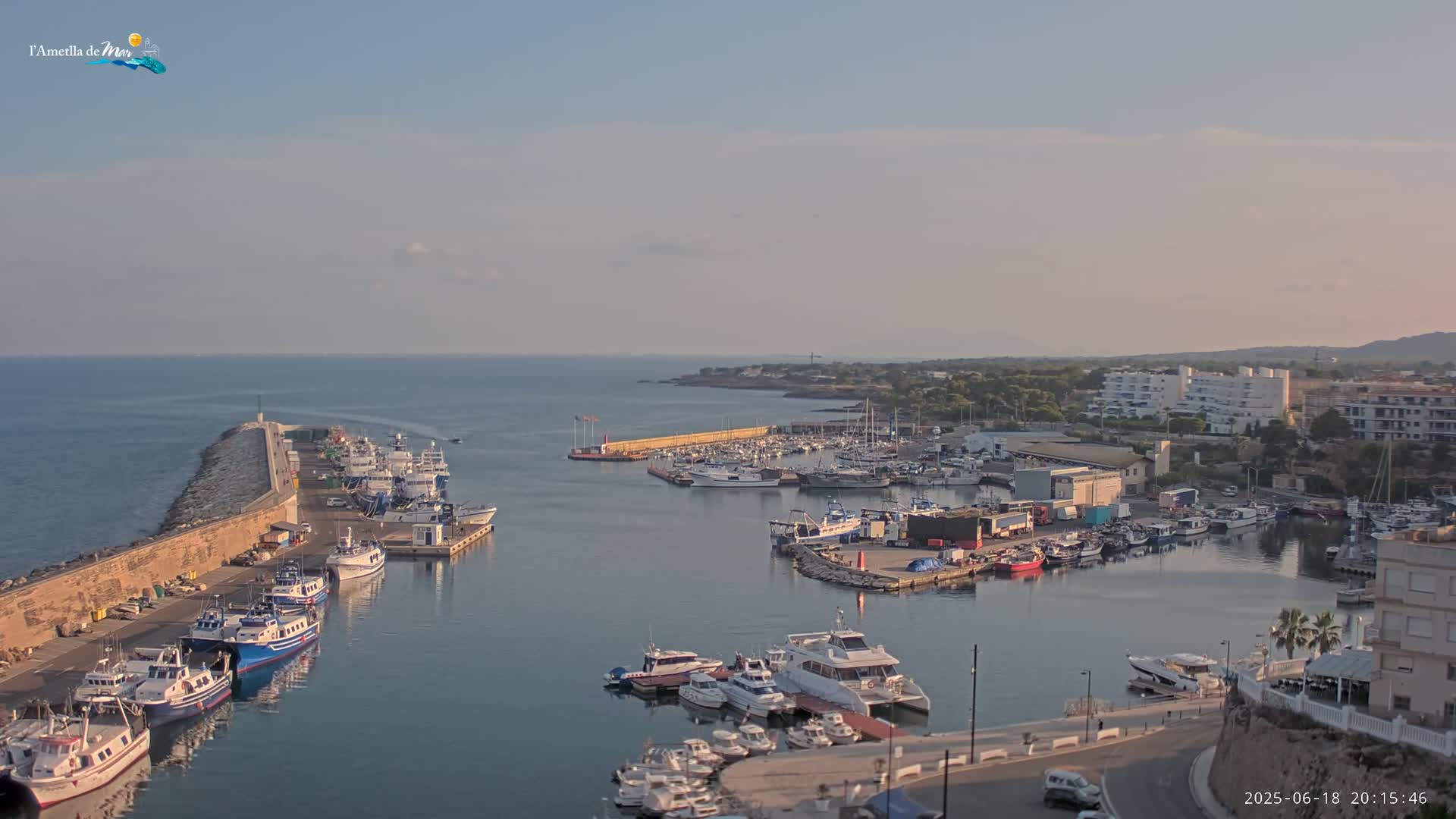 A harbor filled with numerous boats and ships is situated beside a stone wall extending into a calm sea under a clear, slightly hazy sky.