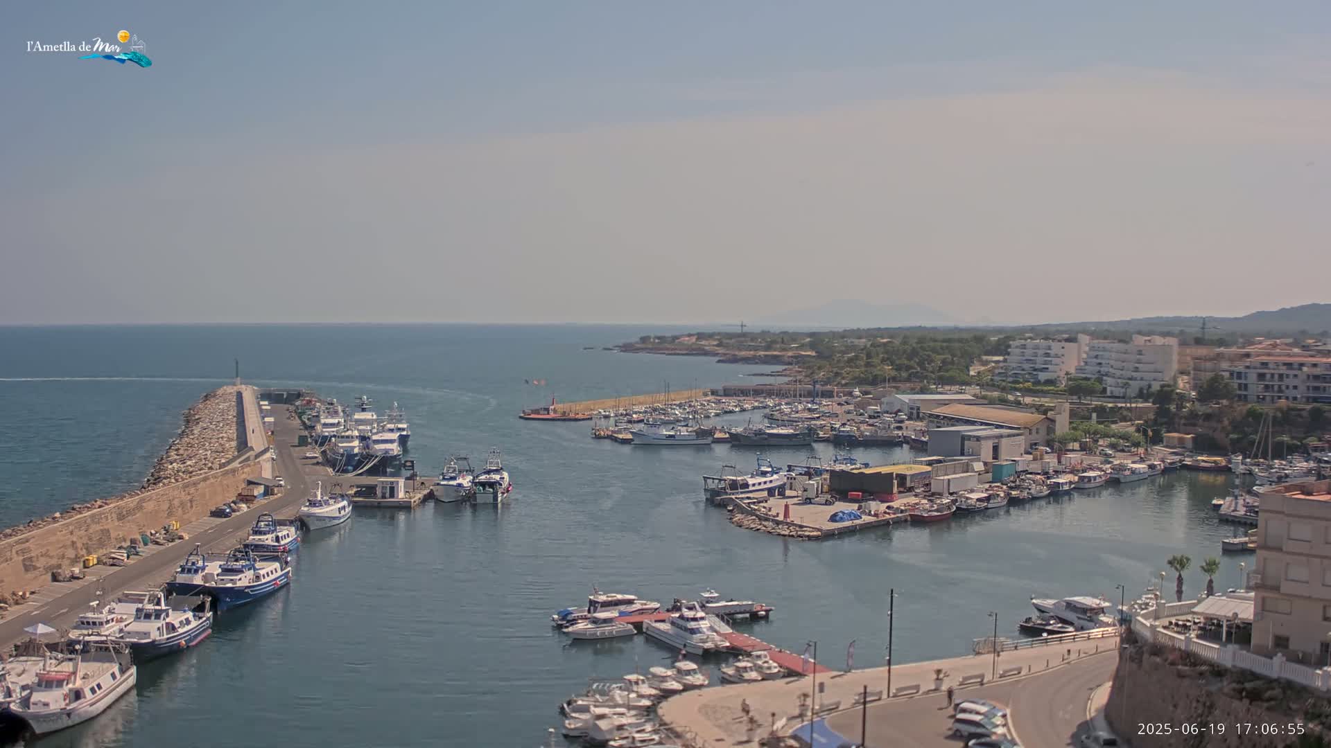 A harbor filled with numerous boats and ships is shown on a sunny day, with calm waters and a distant coastline visible.
