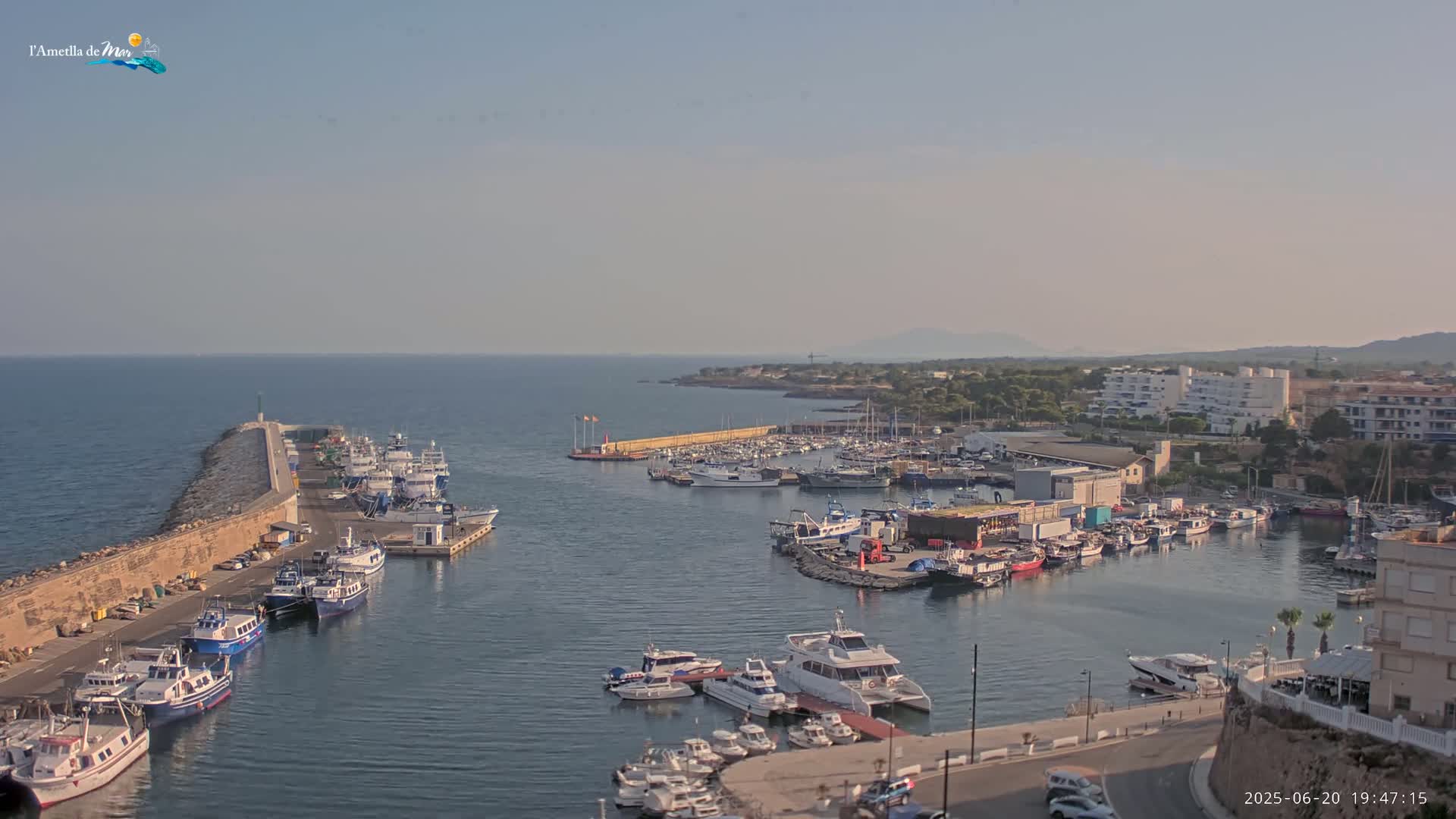 A harbor filled with many boats of various sizes is situated on calm water next to a stone seawall, under a clear, light-blue sky.
