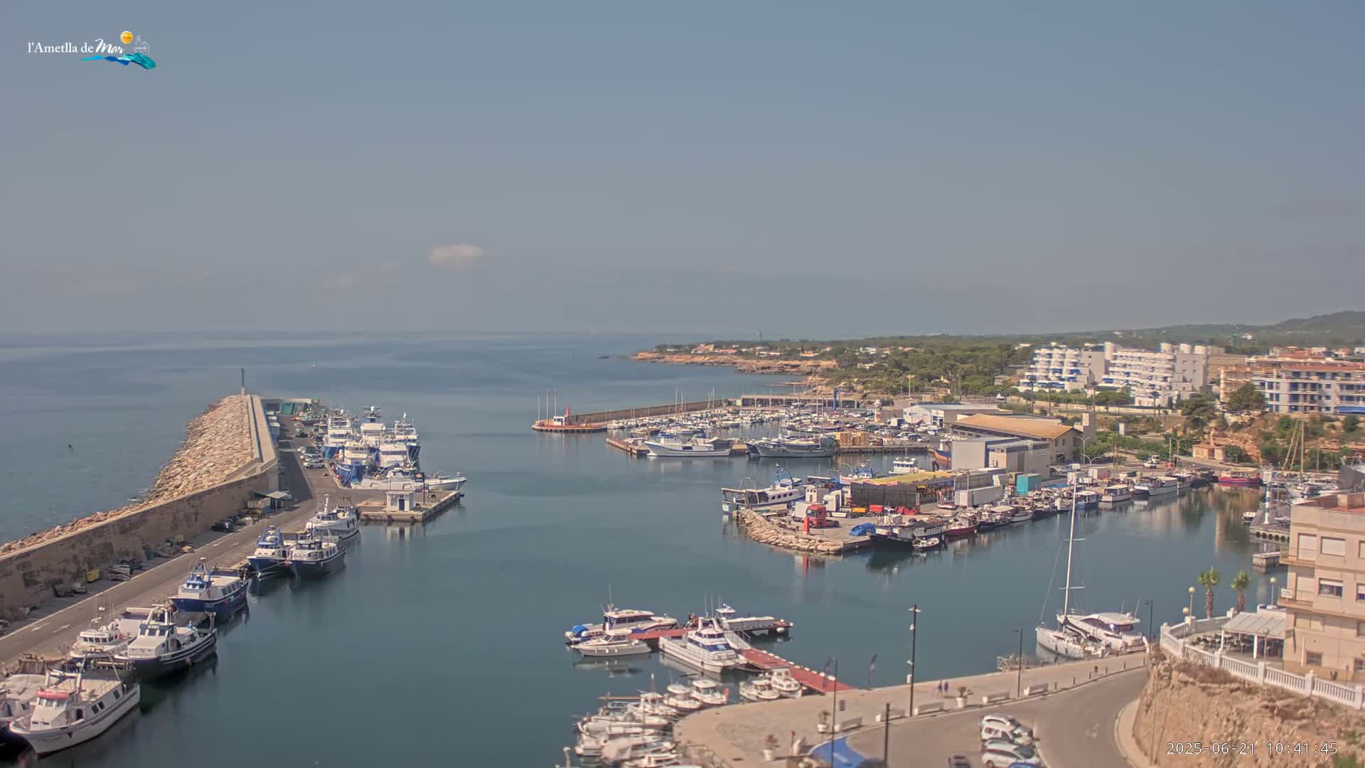 A harbor filled with numerous boats and yachts is nestled along a coastline under a hazy, sunny sky.