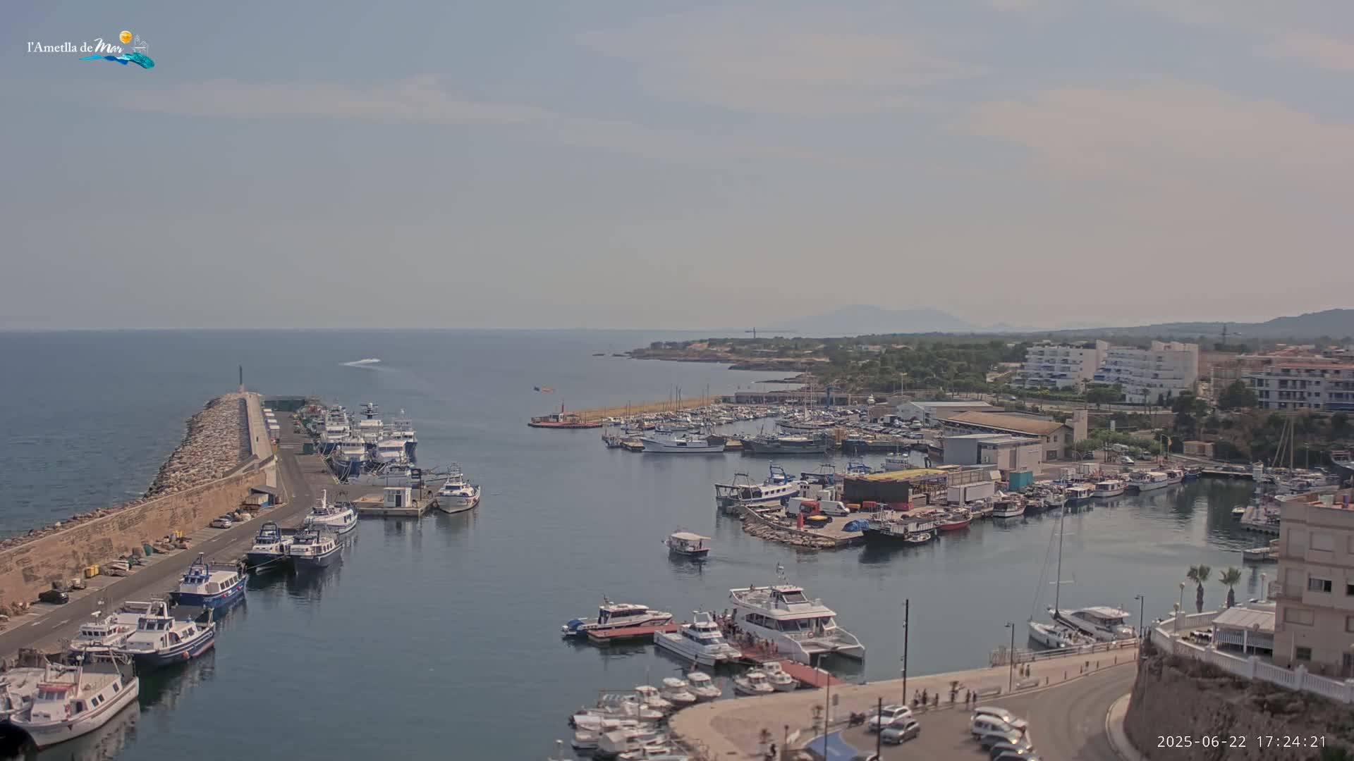 A harbor filled with numerous boats and ships is situated next to a stone wall and overlooks a calm sea under a hazy sky.