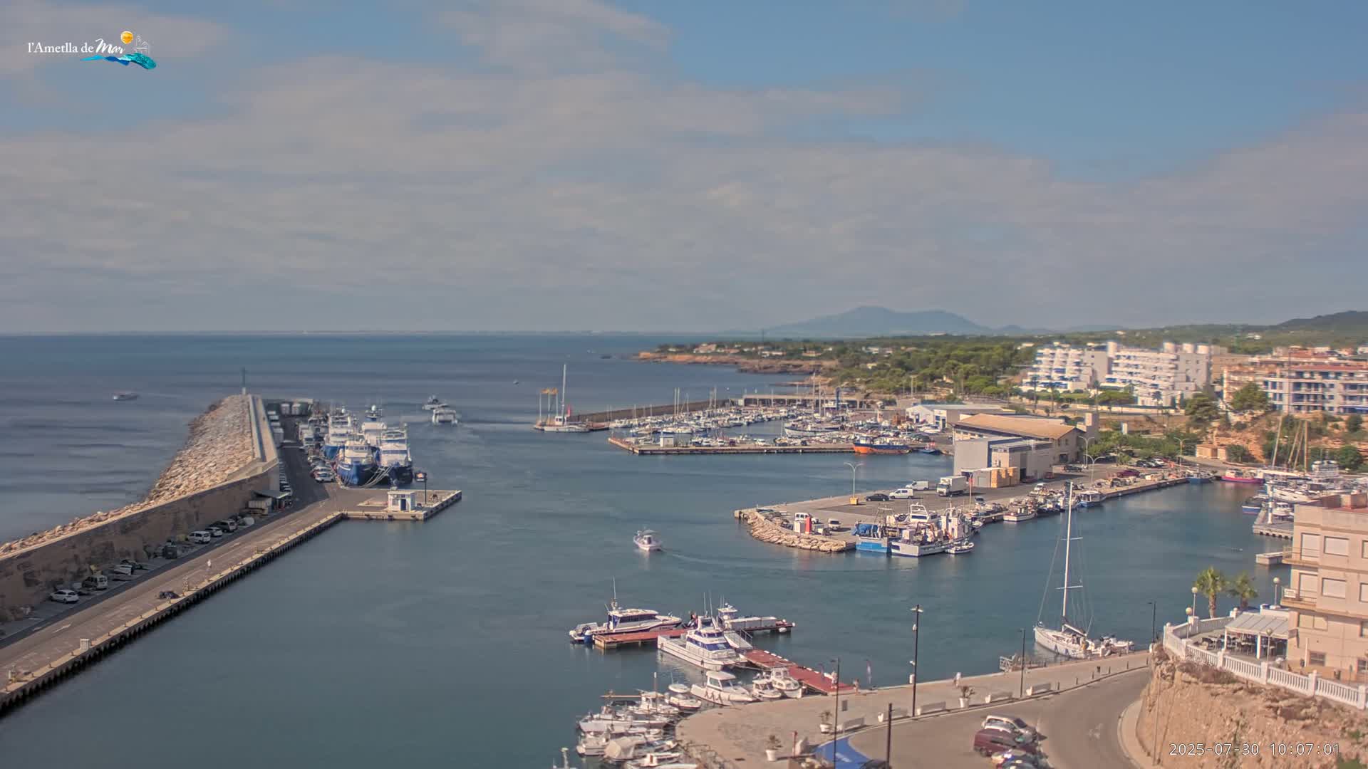 A harbor filled with boats and yachts under a partly cloudy sky, with a breakwater extending into the calm sea.