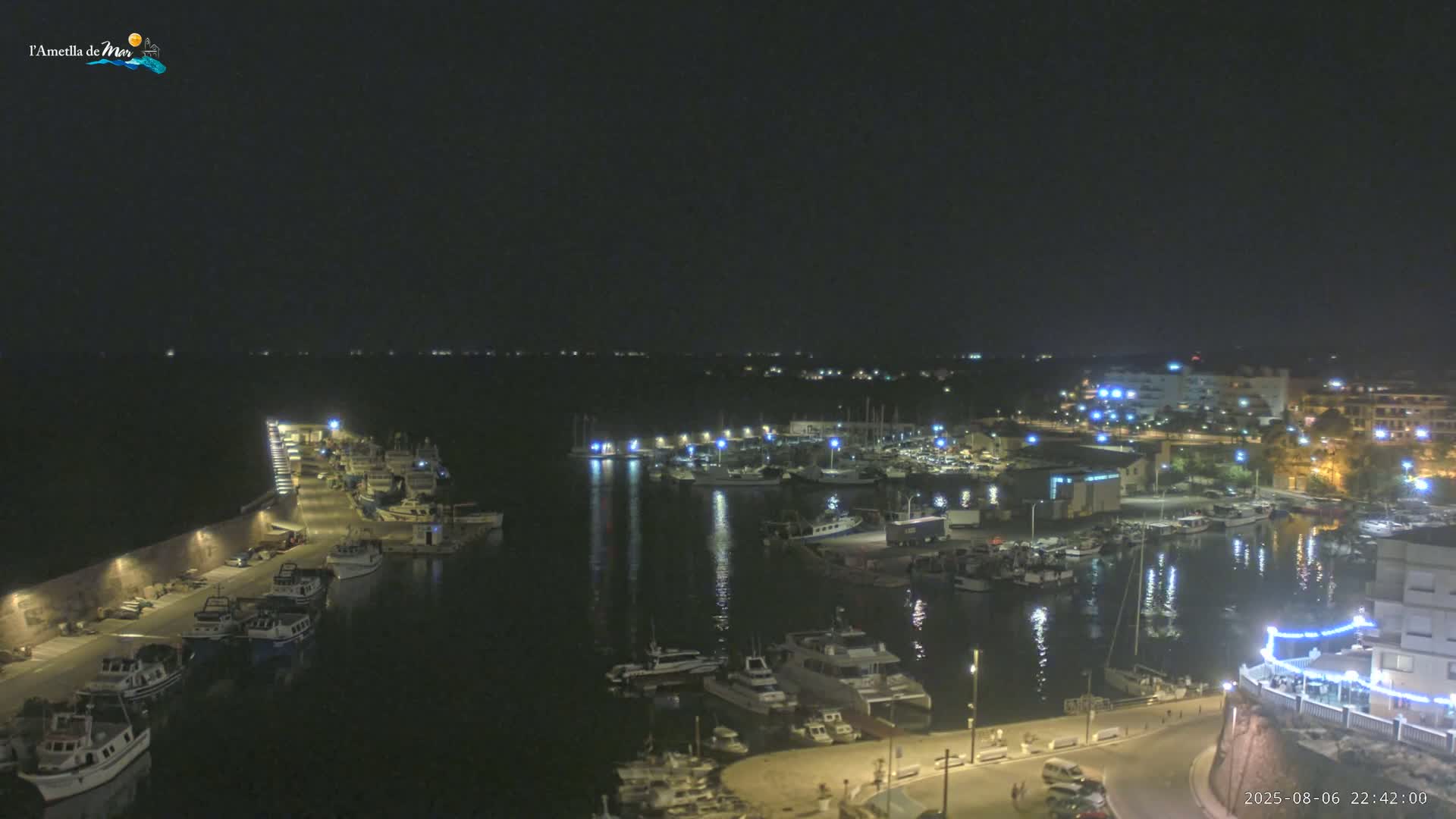 A nighttime aerial view shows a harbor filled with boats, illuminated by lights along the docks and buildings surrounding the calm water.