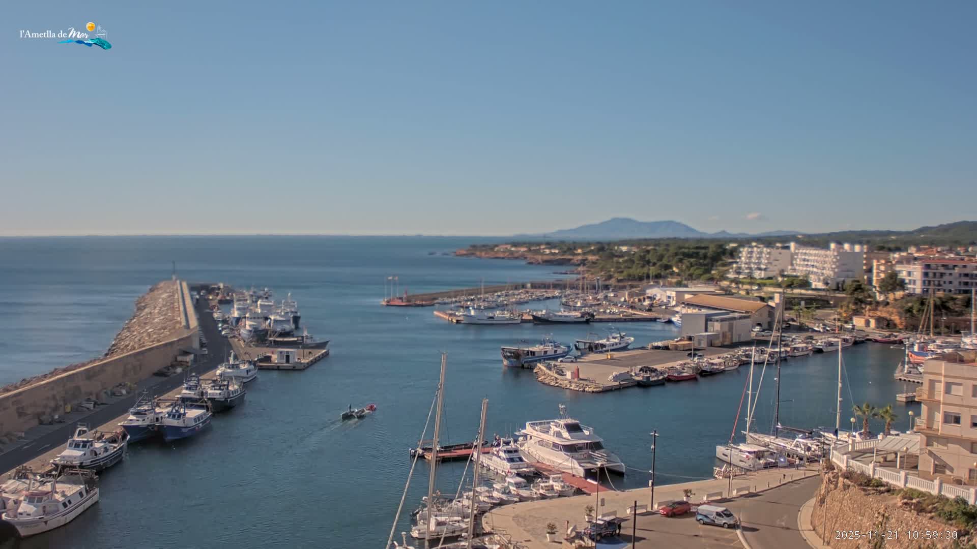 A bustling coastal harbor is seen under a clear blue sky on a sunny day, filled with numerous boats and yachts docked along piers and a long breakwater, with a town and distant mountains visible along the calm, sunlit sea.