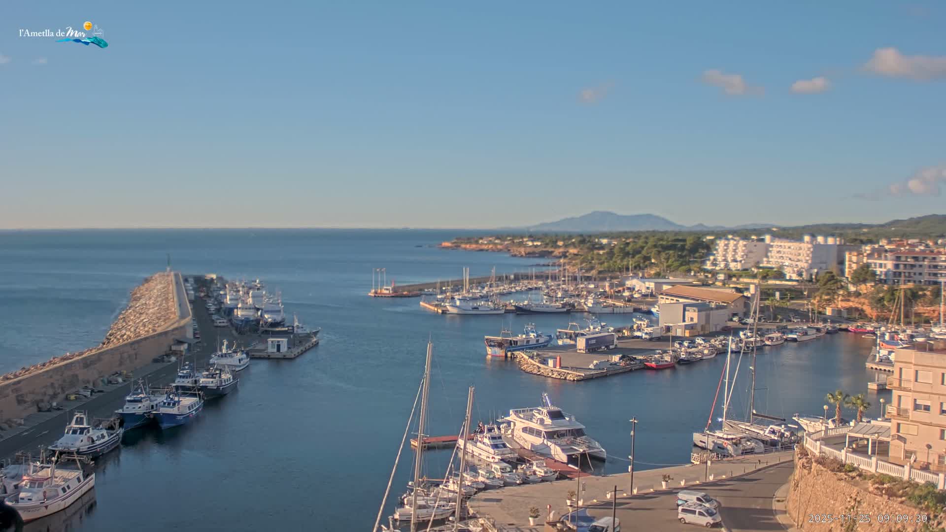 A sunny coastal harbor is depicted with numerous boats docked along piers and a long rocky breakwater, bordered by a town featuring buildings and lush trees, all set against a clear blue sky with scattered clouds and distant mountains.