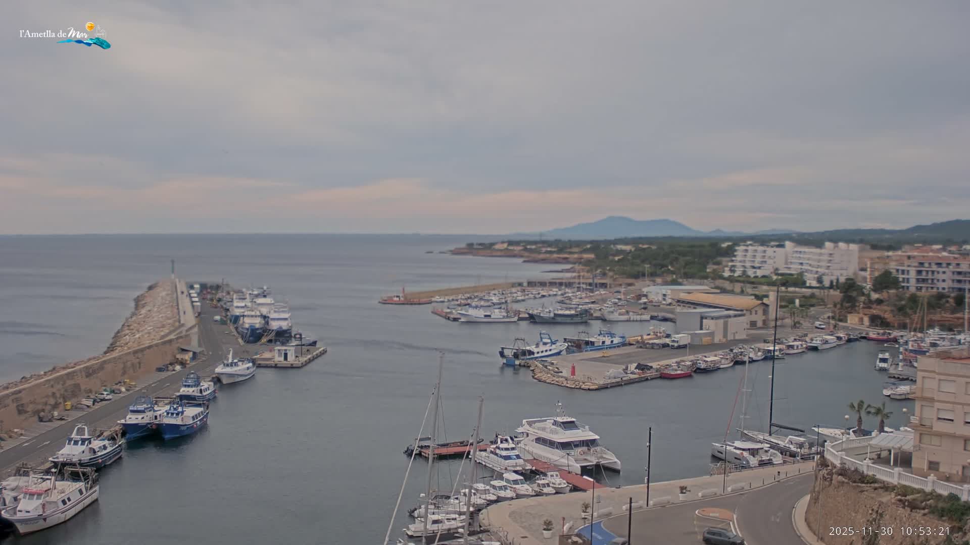 Numerous boats are docked in a calm harbor with a long stone pier extending towards the open sea, where a rugged coastline and distant mountains meet an overcast sky.