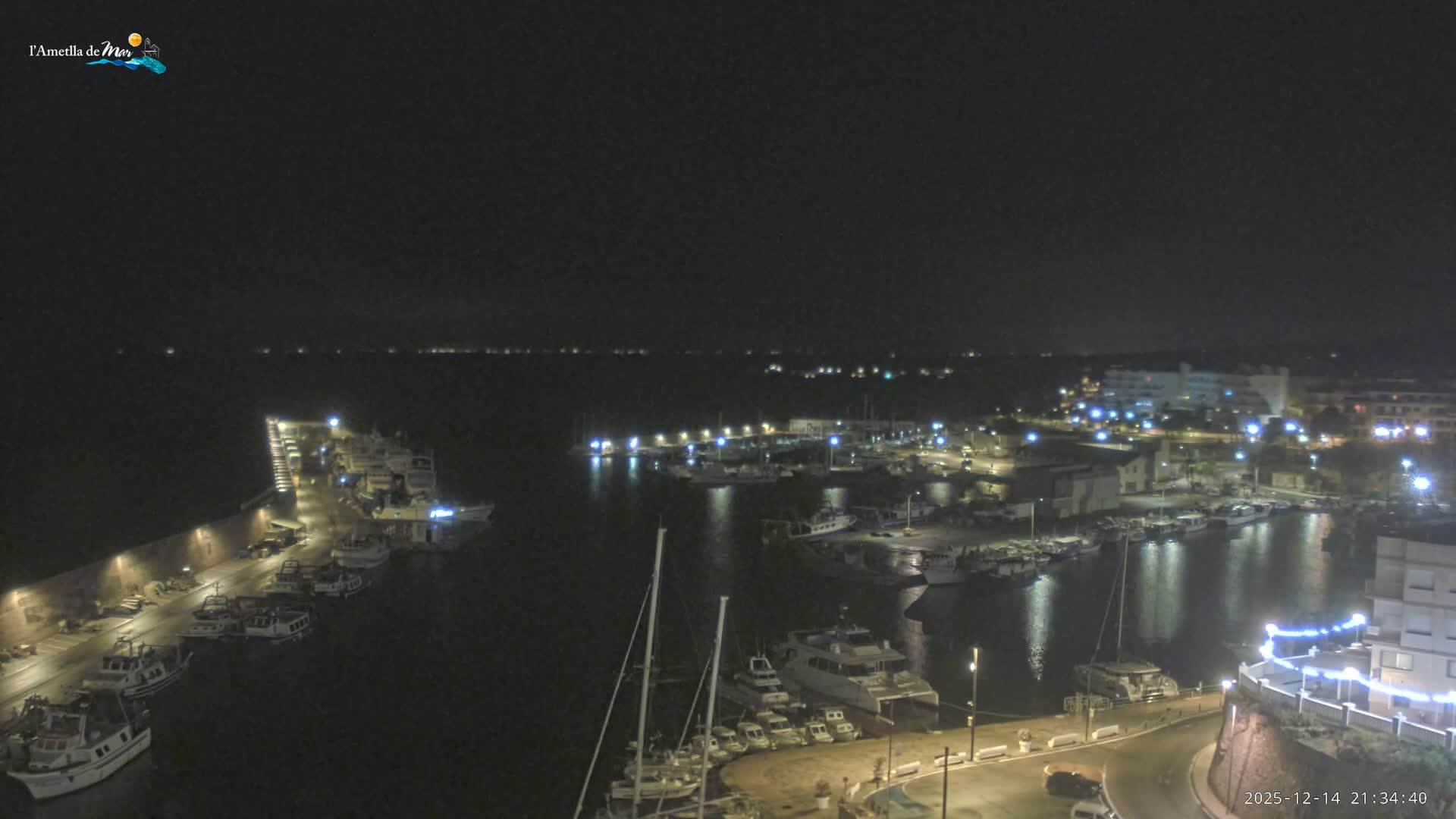 Numerous boats are docked in a calm harbor with a long stone pier extending towards the open sea, where a rugged coastline and distant mountains meet an overcast sky.
