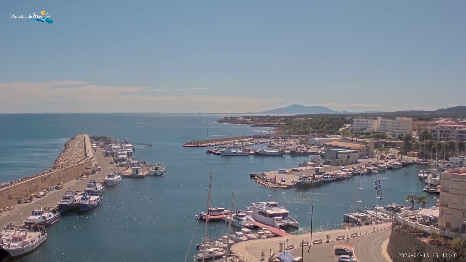 Numerous boats are docked in a calm harbor with a long stone pier extending towards the open sea, where a rugged coastline and distant mountains meet an overcast sky.