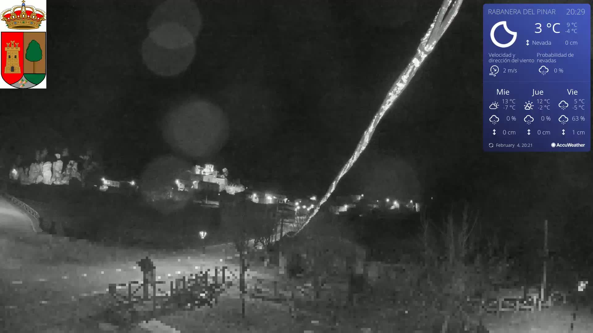 A nighttime view of a snow-dusted town illuminated by lights, under a dark sky with light snowfall and a 3°C temperature.