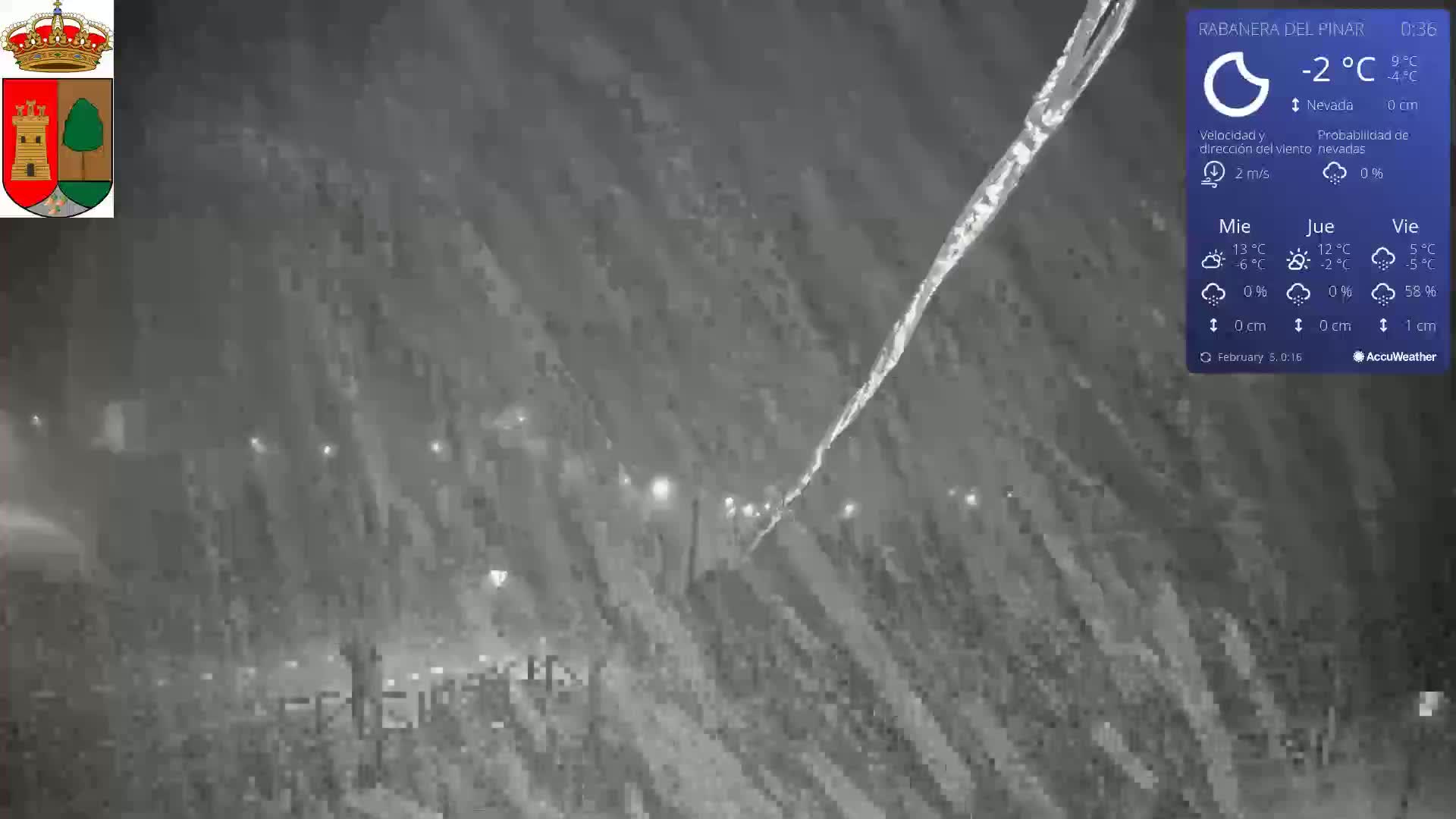 A nighttime scene of a snow-covered landscape under heavy snowfall, with dimly lit streetlights visible in the distance.