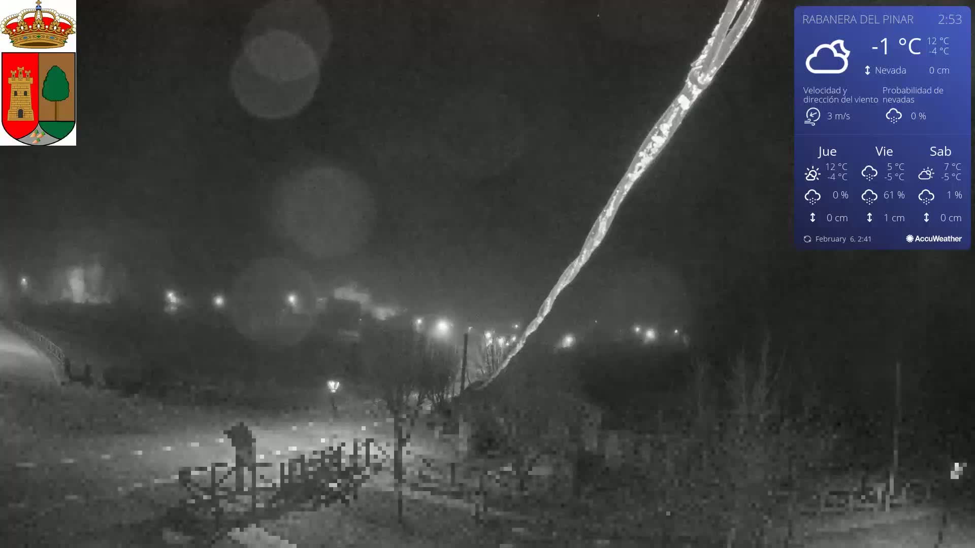 A nighttime, snowy scene shows a long, thin, bright streak in the dark sky above a sparsely lit area with some trees and a fence.