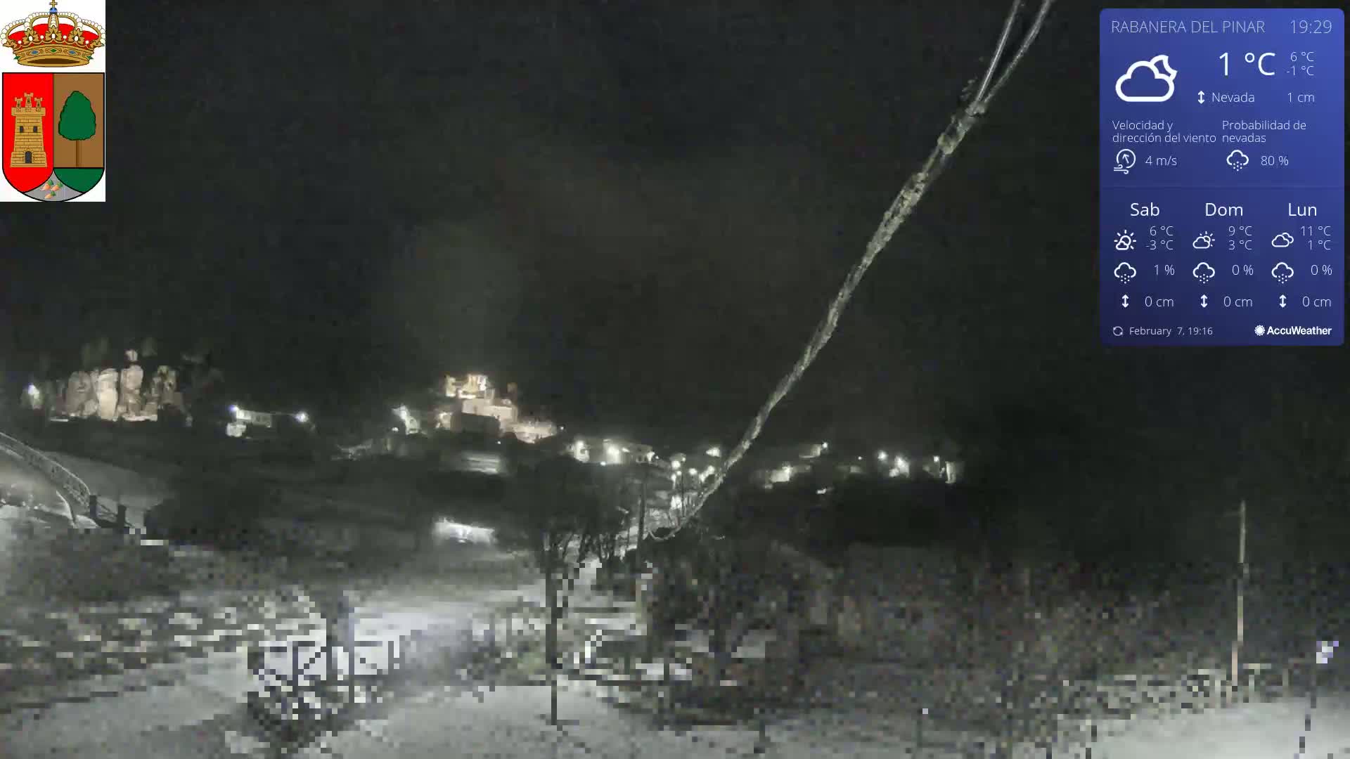 A nighttime, snowy scene shows a village with dimly lit buildings and a few cars under a dark sky.