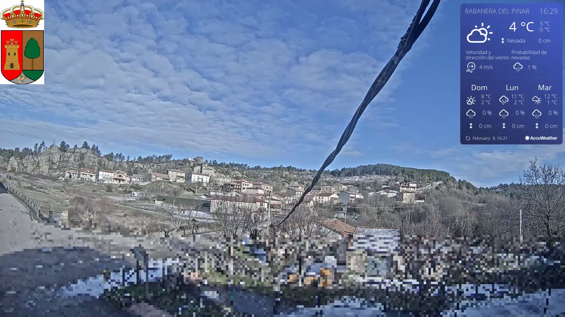 A partly cloudy sky over a small town nestled in a hilly landscape with some snow on the ground.