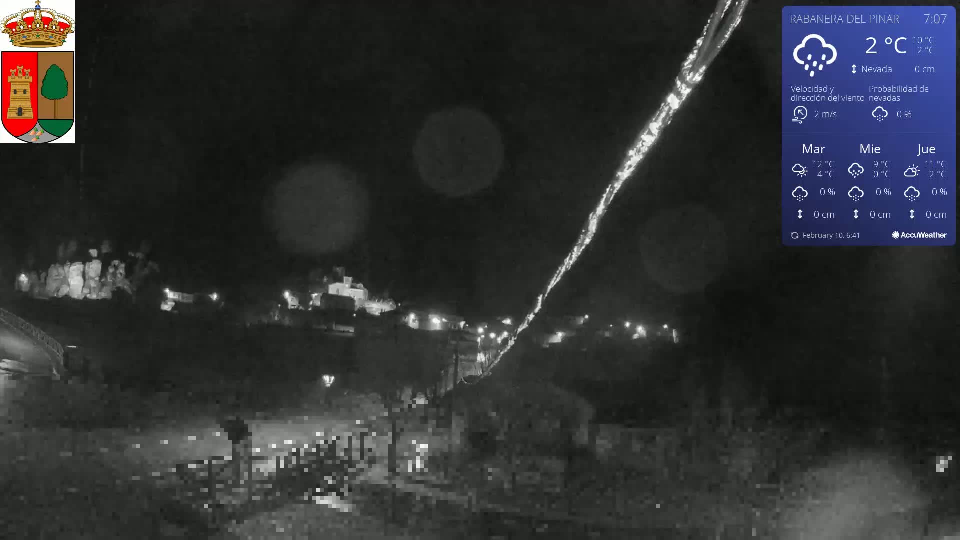 A nighttime view of a snow-covered town, illuminated by scattered lights under a lightly snowing sky.