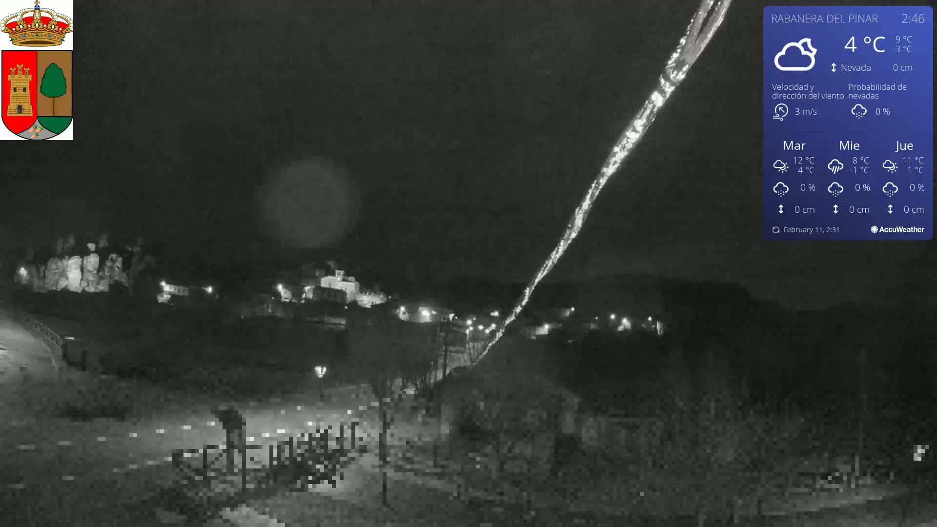 A nighttime, low-light view of a town illuminated by scattered lights, with a bright streak across the dark sky under mostly cloudy conditions and a temperature of 4°C.